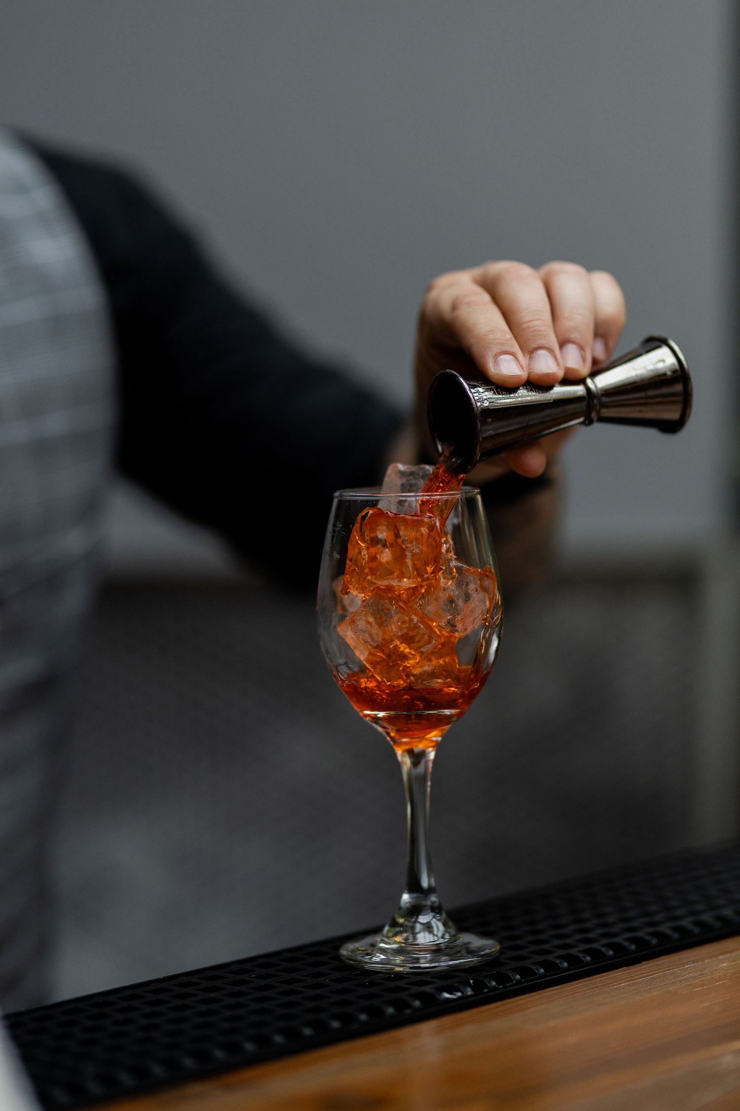 A hand pours liquid from a jigger into an orange cocktail in a wine glass on a black bar mat.