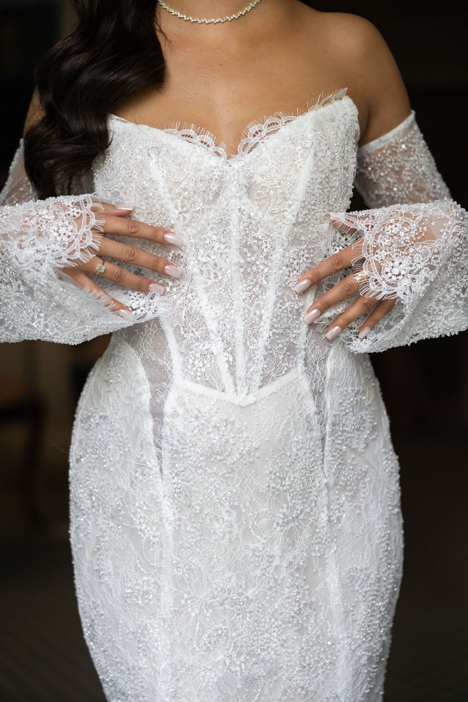 Bride in white lace off-shoulder corset wedding gown with bell sleeves and intricate beading, hands posed on waist.