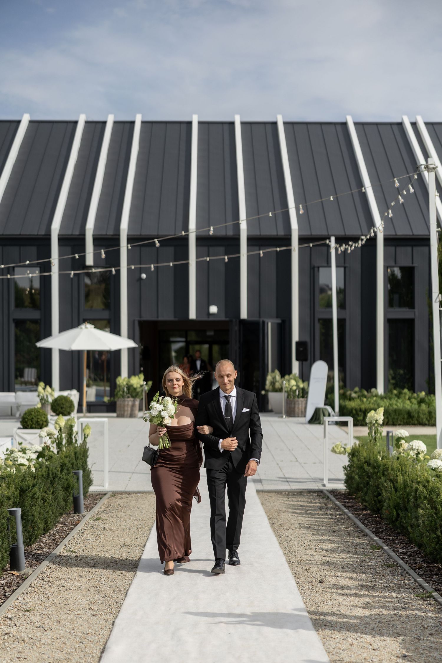 Elegant couple walking down a white pathway outside a modern black wedding venue with string lights and white floral arrangements.