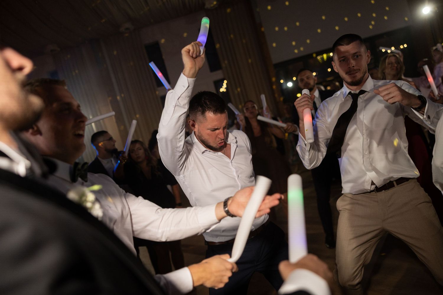 Men dancing energetically at a wedding reception, holding colorful glow sticks under warm venue lighting.