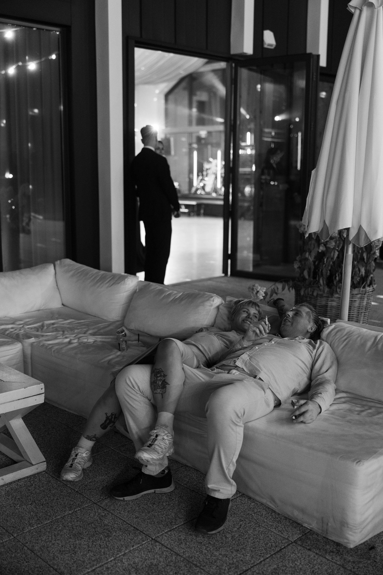 Black and white photo of two people relaxing on a white sofa outdoors at night, with a man standing in background.