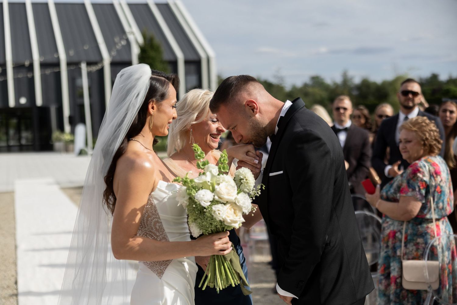 Bride in white gown and groom in tuxedo share a tender moment with a guest holding white bouquet at outdoor wedding ceremony.