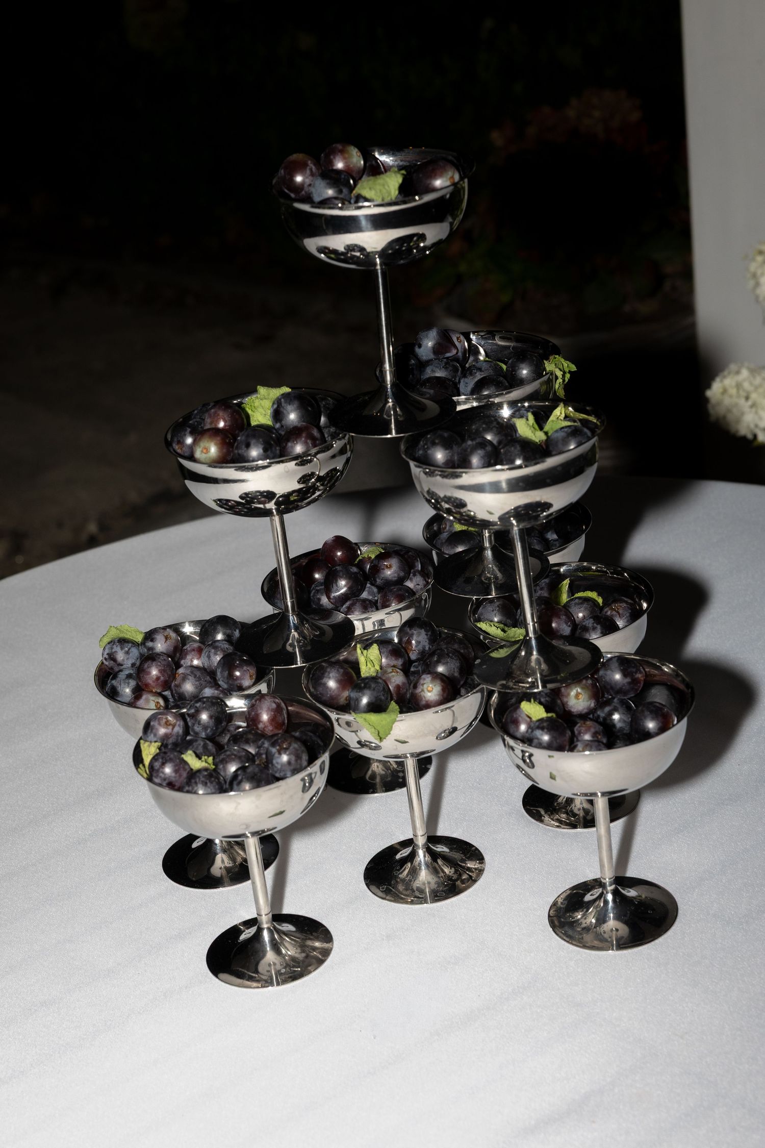 Elegant tiered display of silver goblets filled with fresh dark grapes and mint leaves on a white tablecloth.