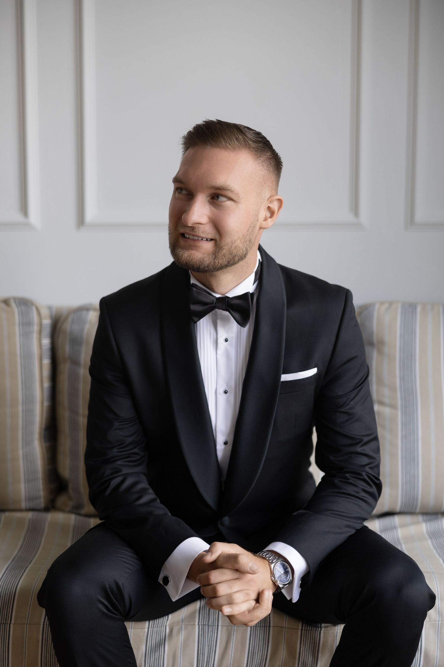 Smiling man in black tuxedo with bow tie sits on striped sofa, hands clasped, wearing a watch.