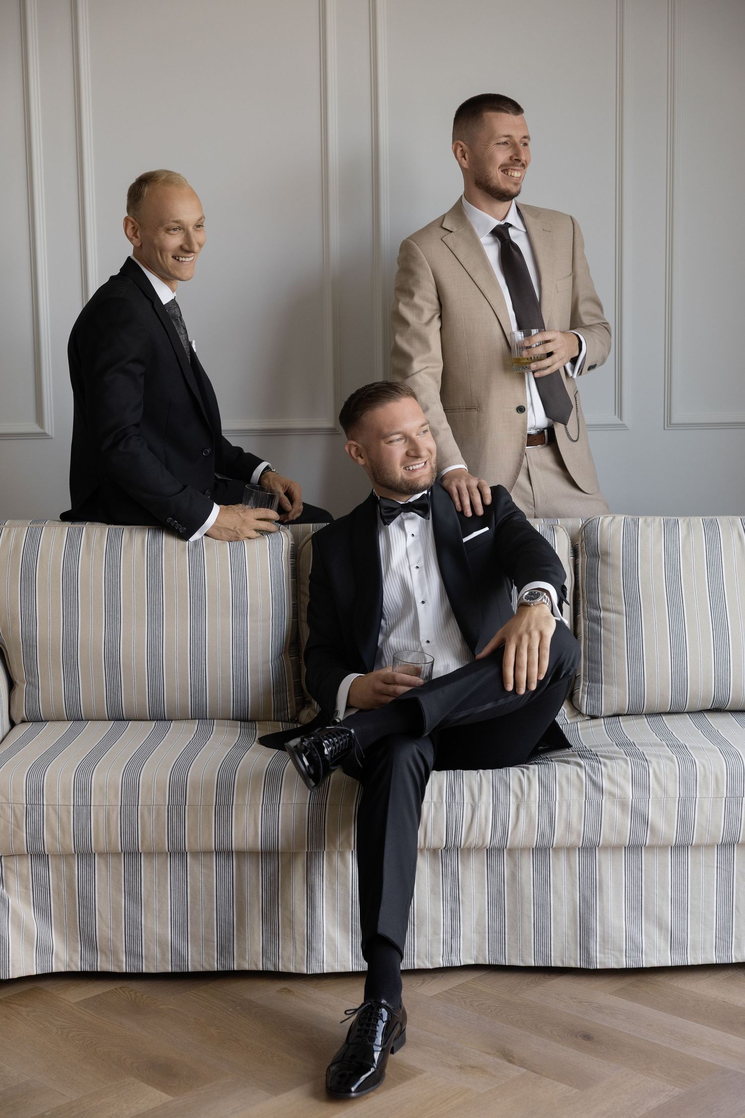 Three well-dressed men in suits smiling and relaxing on a striped sofa in an elegant room.