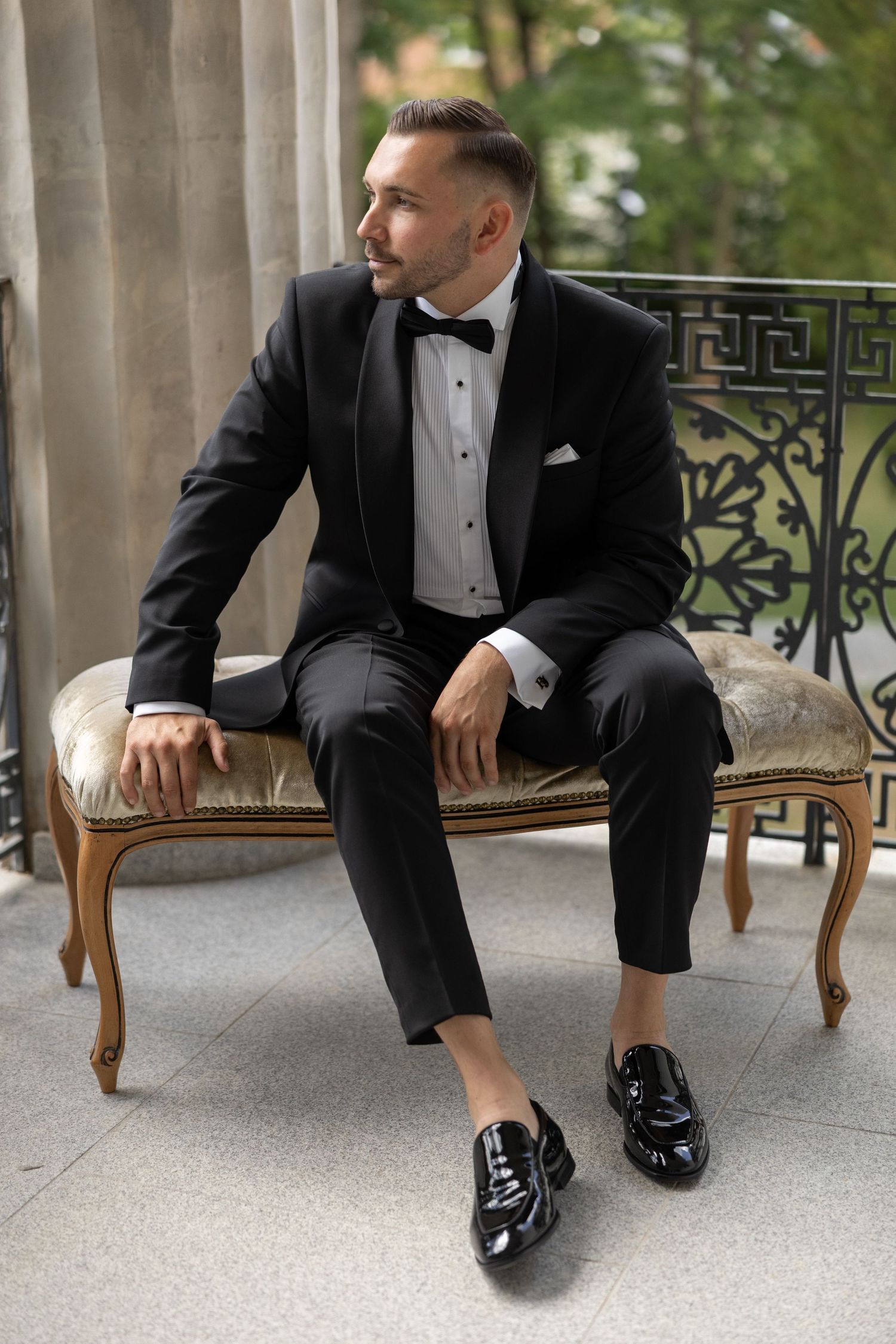Man in black tuxedo with bow tie seated on vintage bench, wearing patent leather loafers on outdoor balcony.