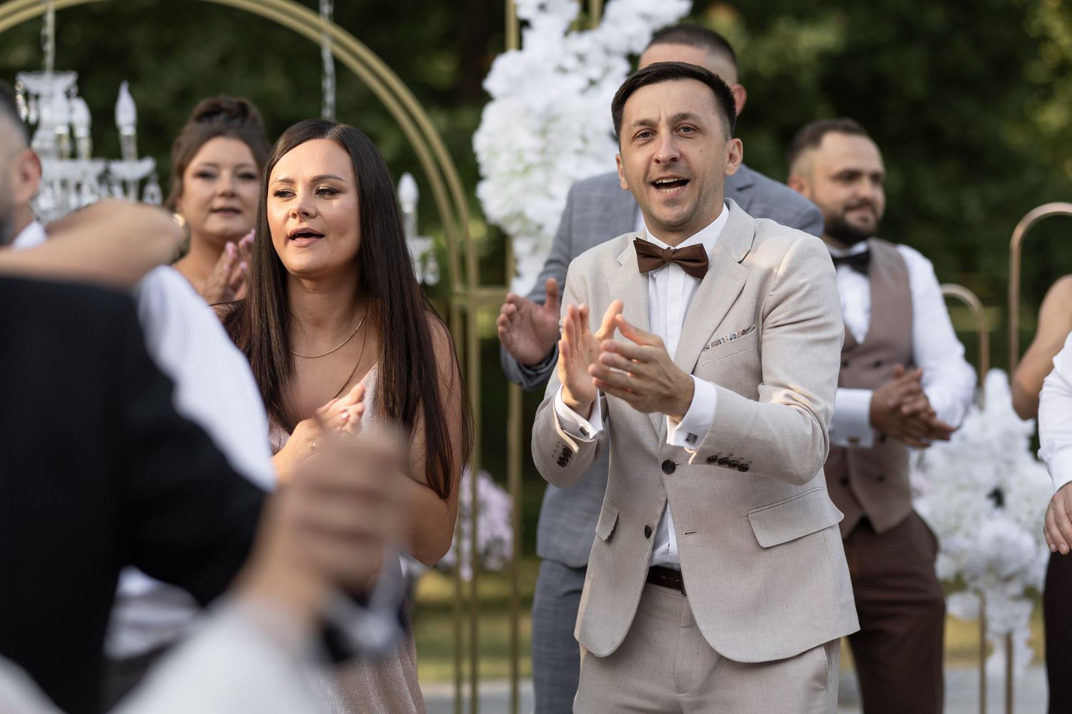 Guests clapping and celebrating at an outdoor wedding ceremony with floral arch decorations in background.