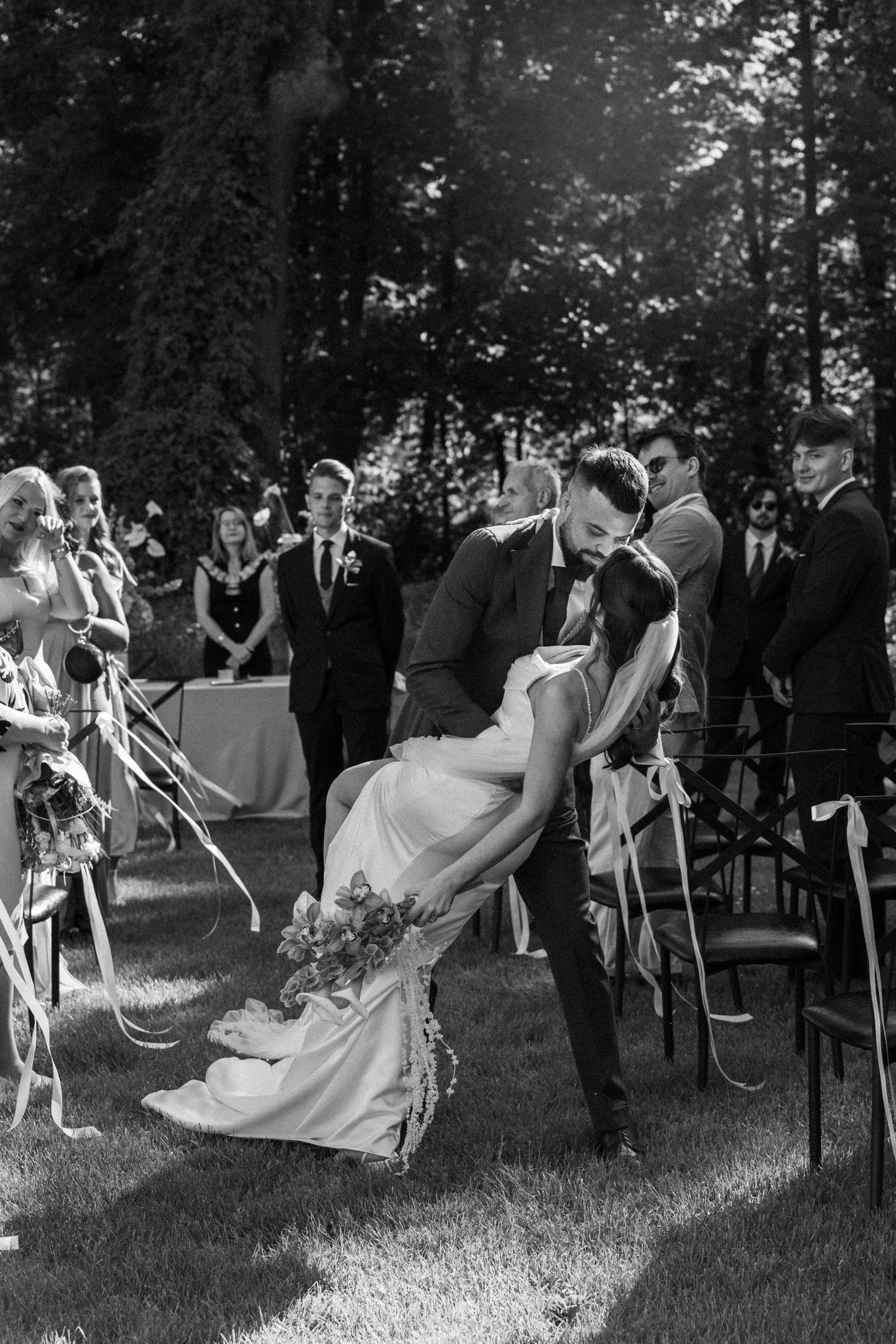 A romantic black and white wedding photo of a couple sharing an intimate dip kiss during their outdoor ceremony.