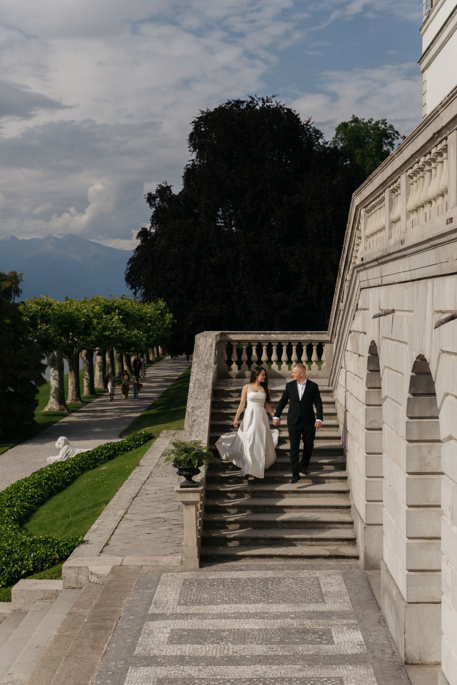 Bride and groom walk up elegant stone stairs at a grand villa with manicured gardens and mountain views.