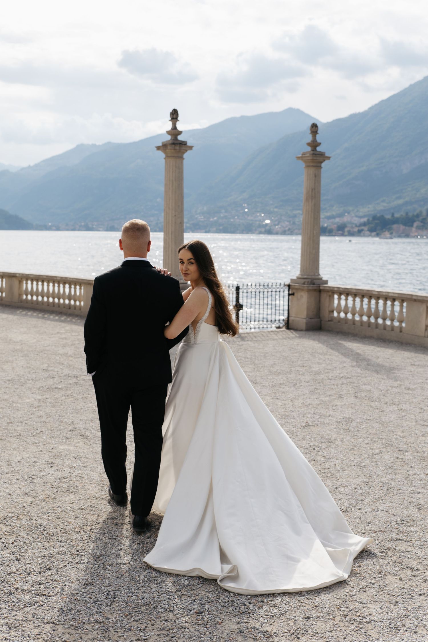 Bride in white gown and groom in black tuxedo walk on terrace overlooking Lake Como with mountains backdrop.