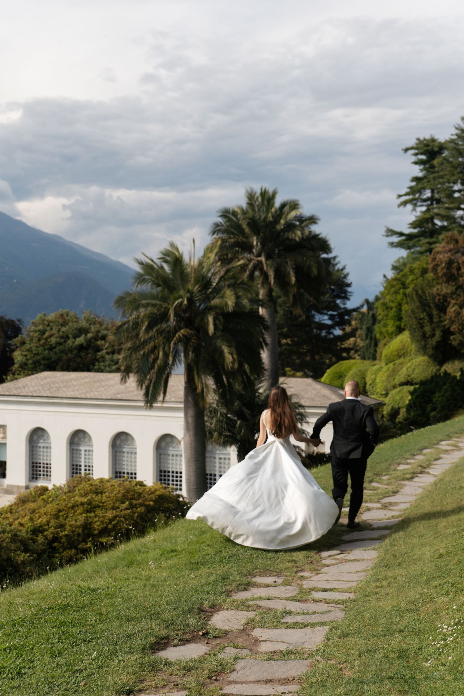 Bride and groom walking hand in hand on a stone path toward an elegant villa surrounded by palm trees and mountains.