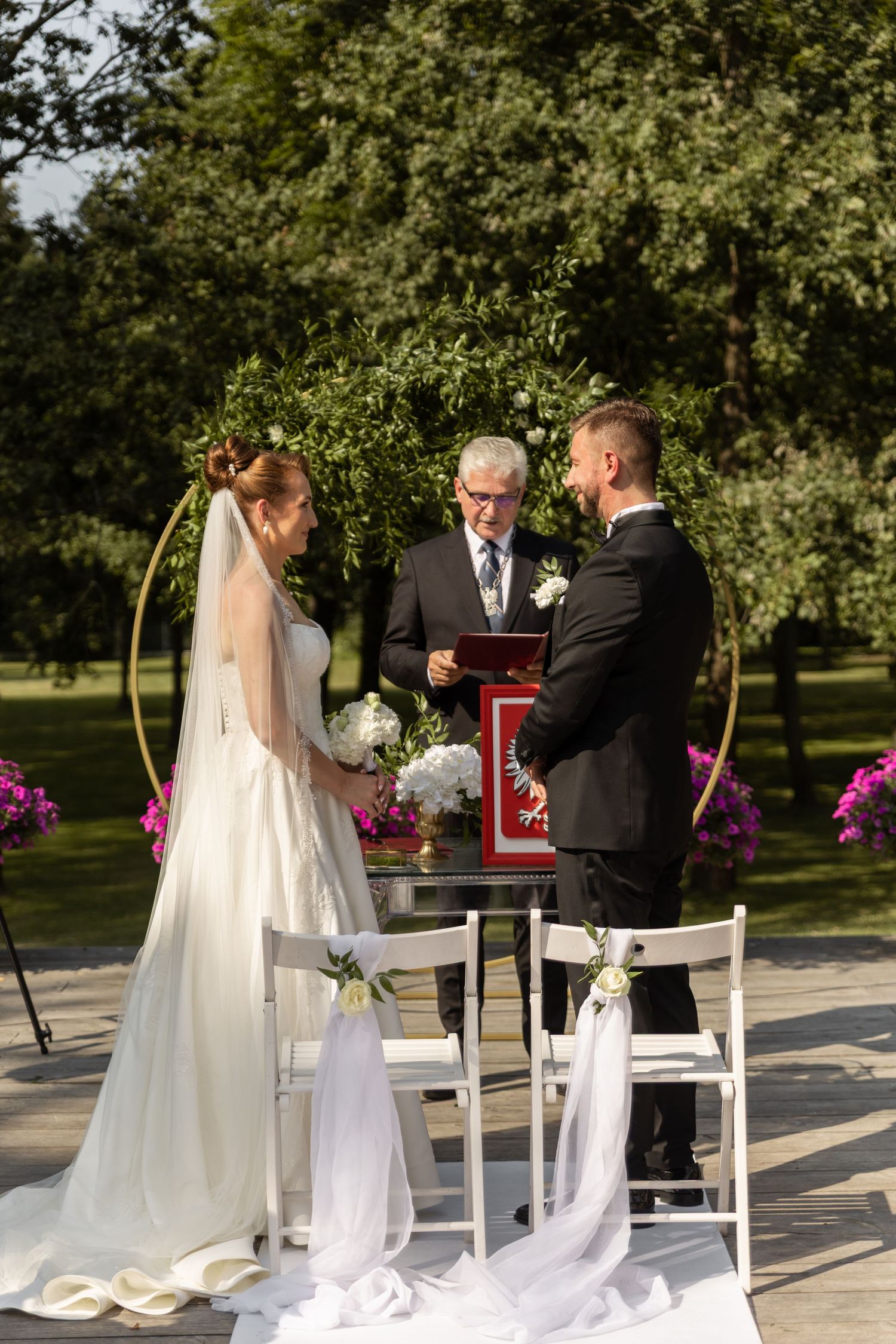 A wedding ceremony taking place outdoors with white chairs and floral decorations in a garden setting.