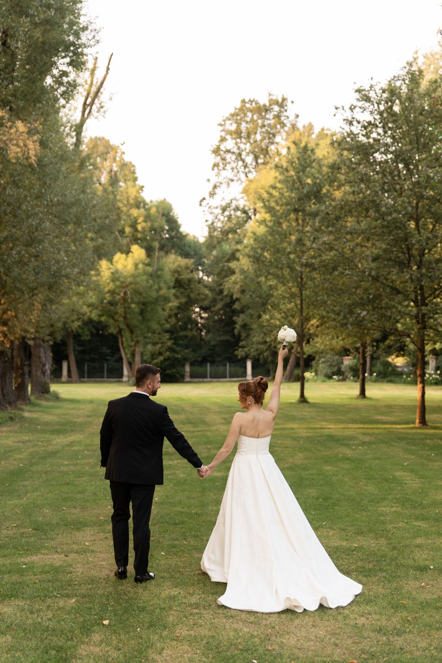 A couple in formal wedding attire walks together across a sunlit grassy lawn surrounded by trees at golden hour.