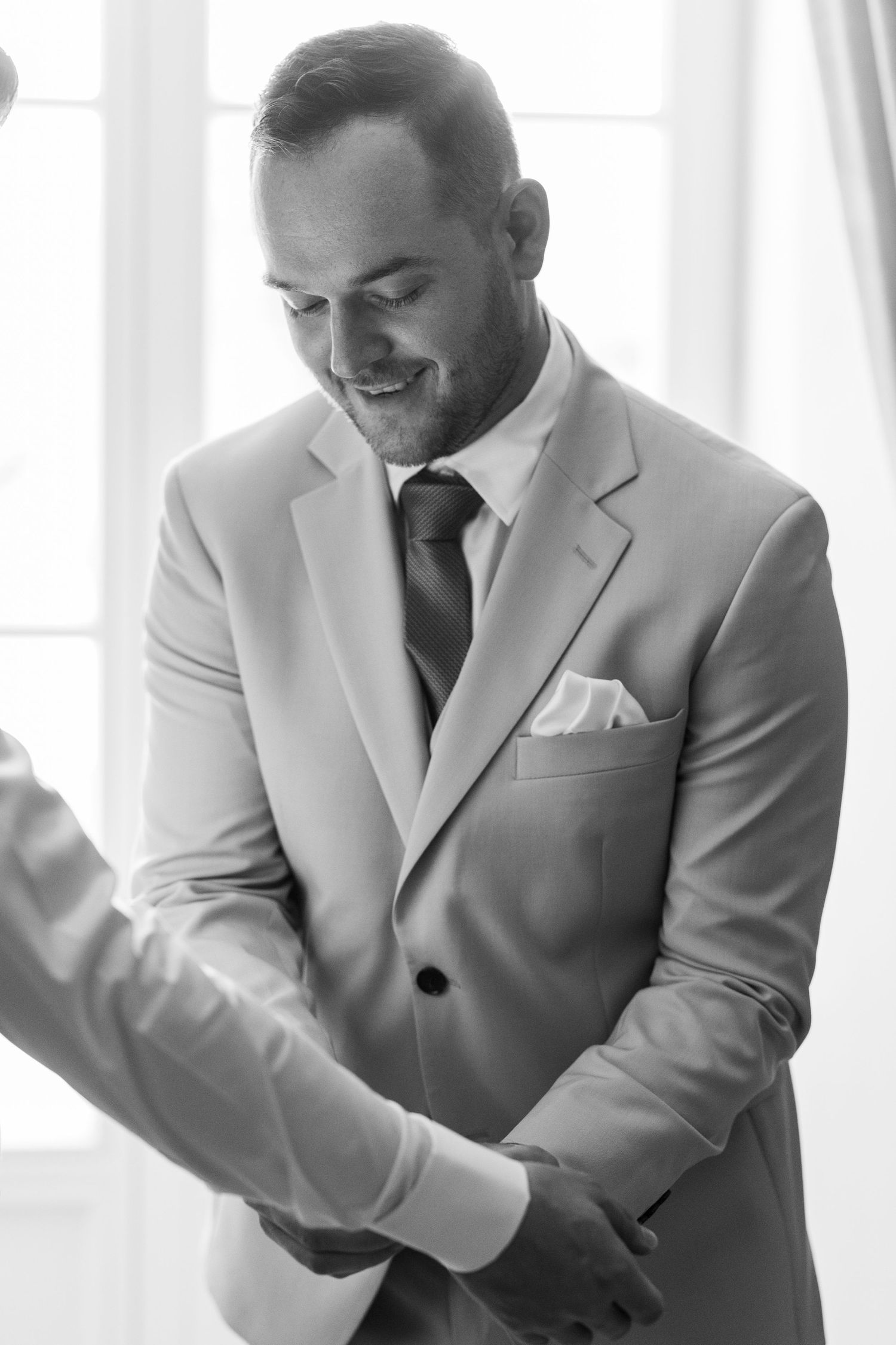 A person in a light gray suit smiles and adjusts their sleeve cuffs in a bright sunlit room.