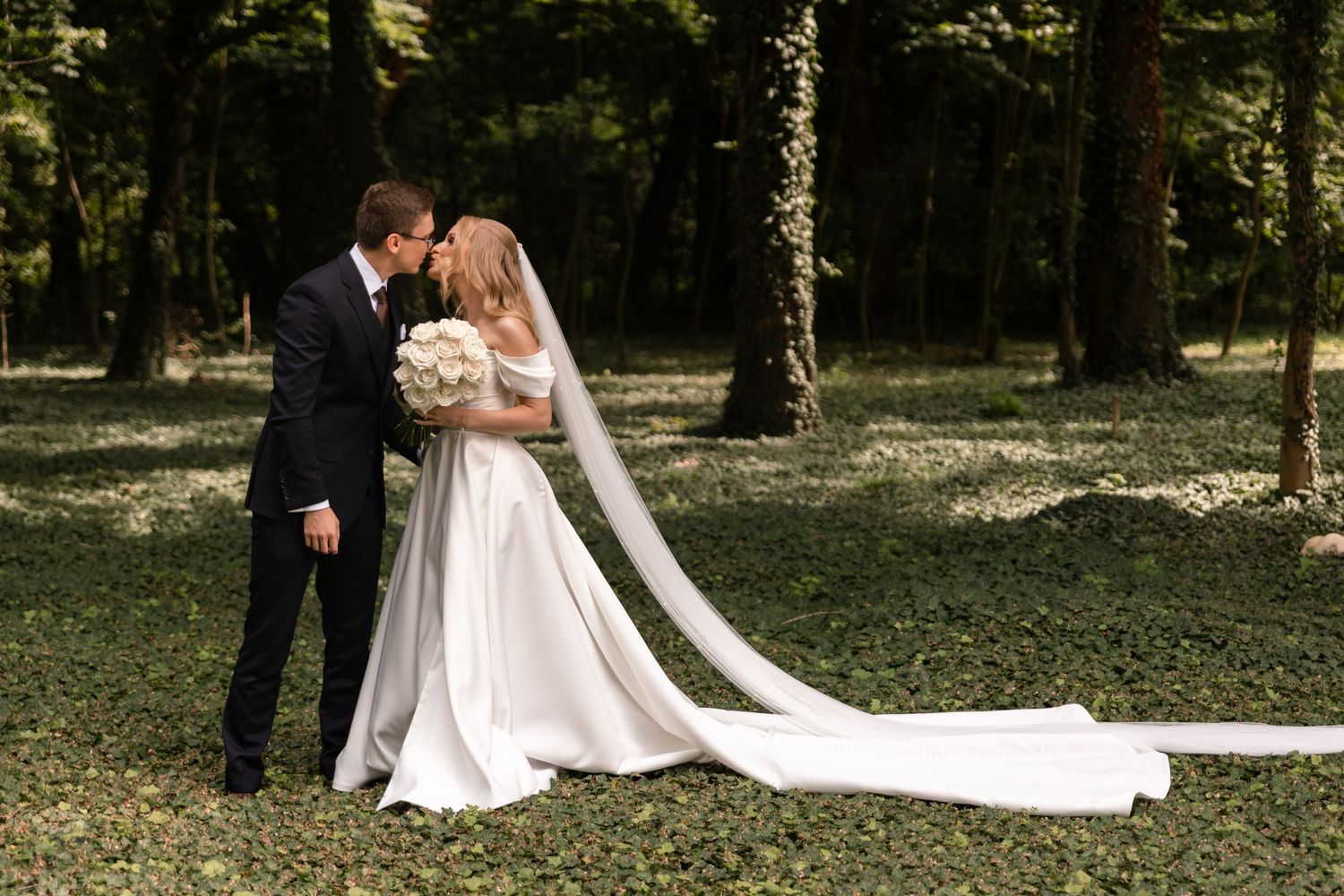 A couple shares romantic moments in a wooded outdoor setting wearing formal wedding attire.