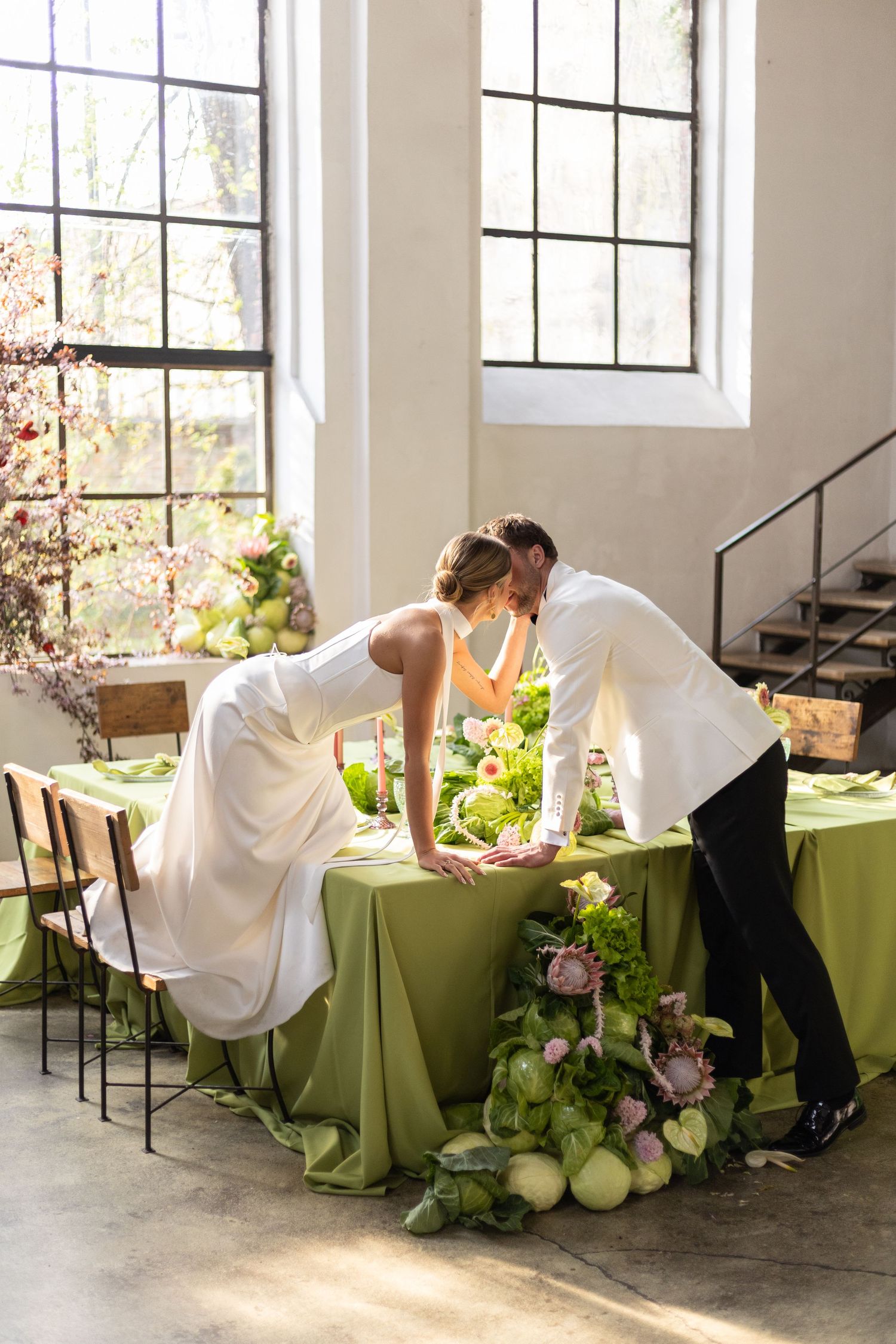 Bride and groom sharing a kiss over a green-draped wedding table adorned with lush floral arrangements indoors.