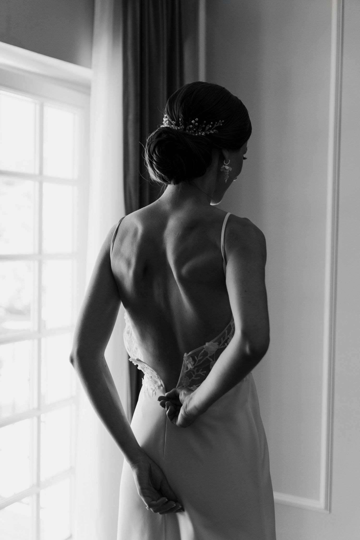 Black and white elegant wedding photo showing back view of bride in sleek dress zipping up by window.