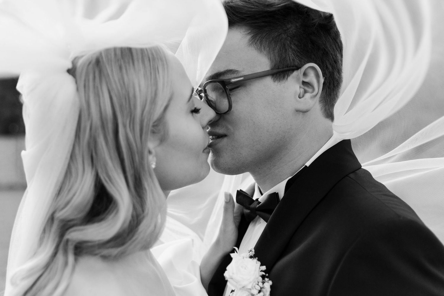 A romantic black and white portrait of newlyweds sharing a tender kiss with flowing veil in the background.