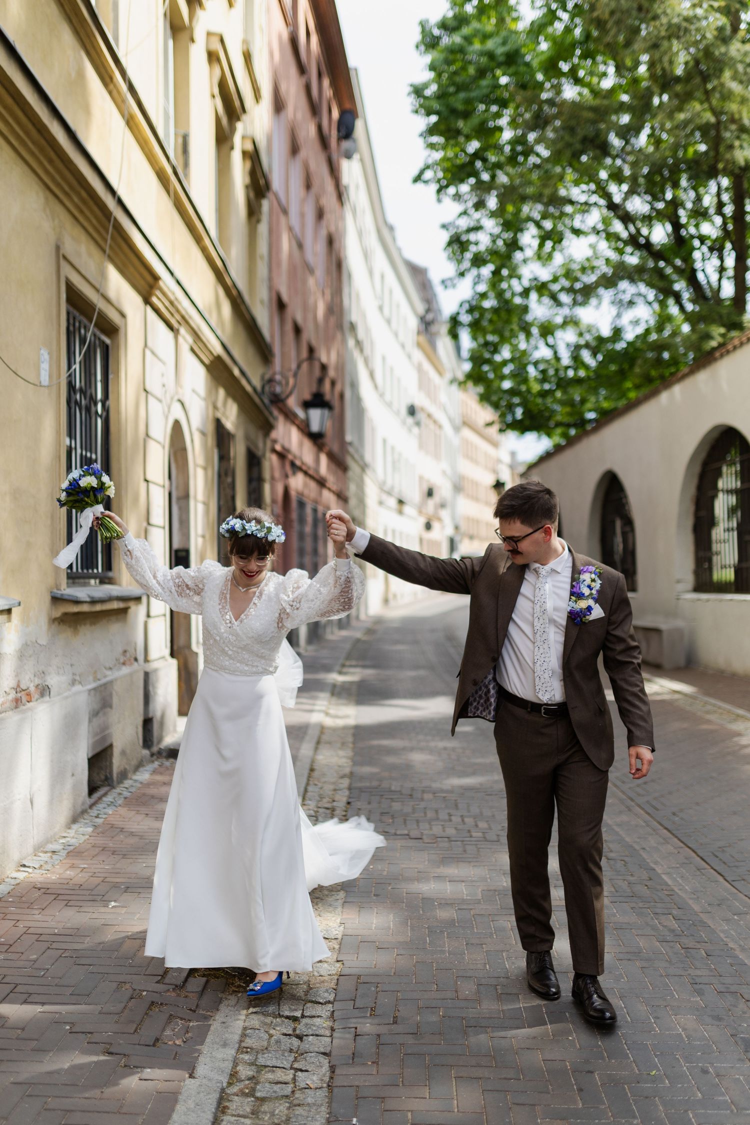 A couple in wedding attire dance together on a charming European cobblestone street lined with historic buildings.
