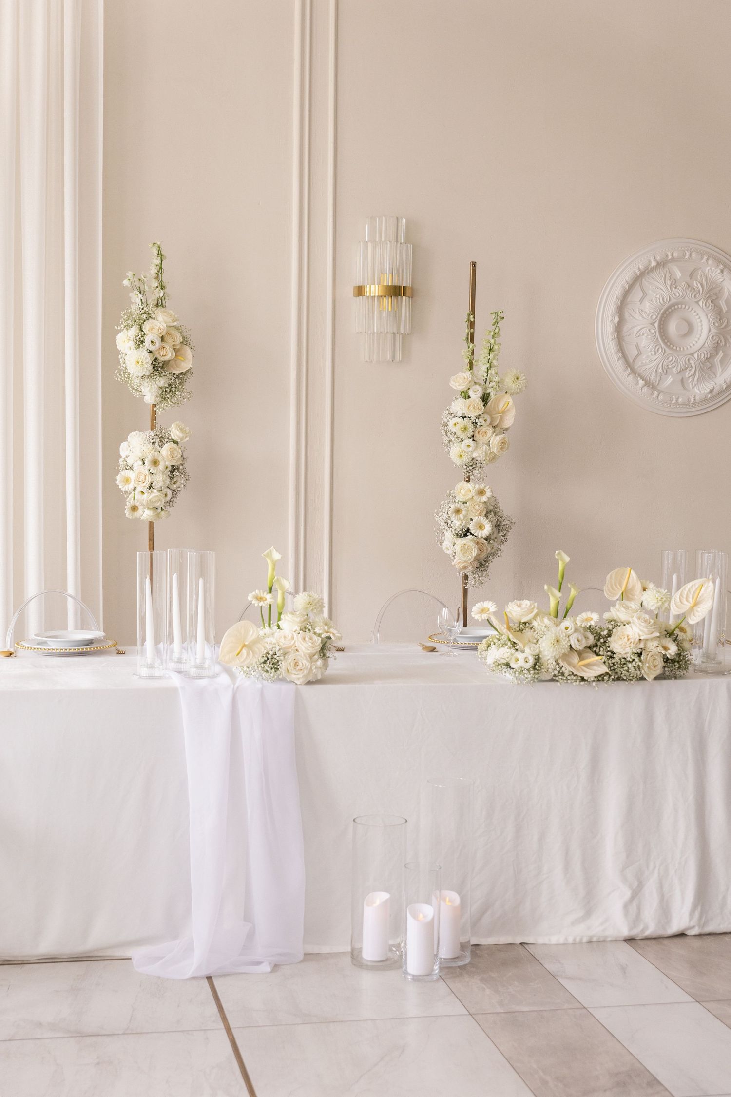 Elegant white floral arrangements on tables with delicate white linens in a light-filled room with ornate wall details.