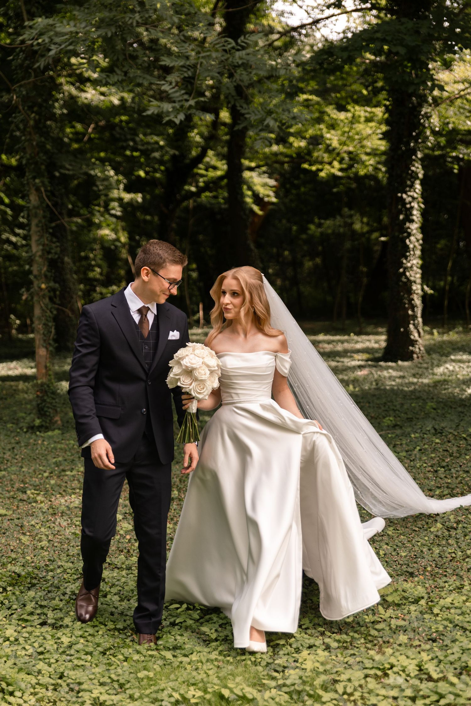 A couple in wedding attire walks together through a lush wooded area on their special day.
