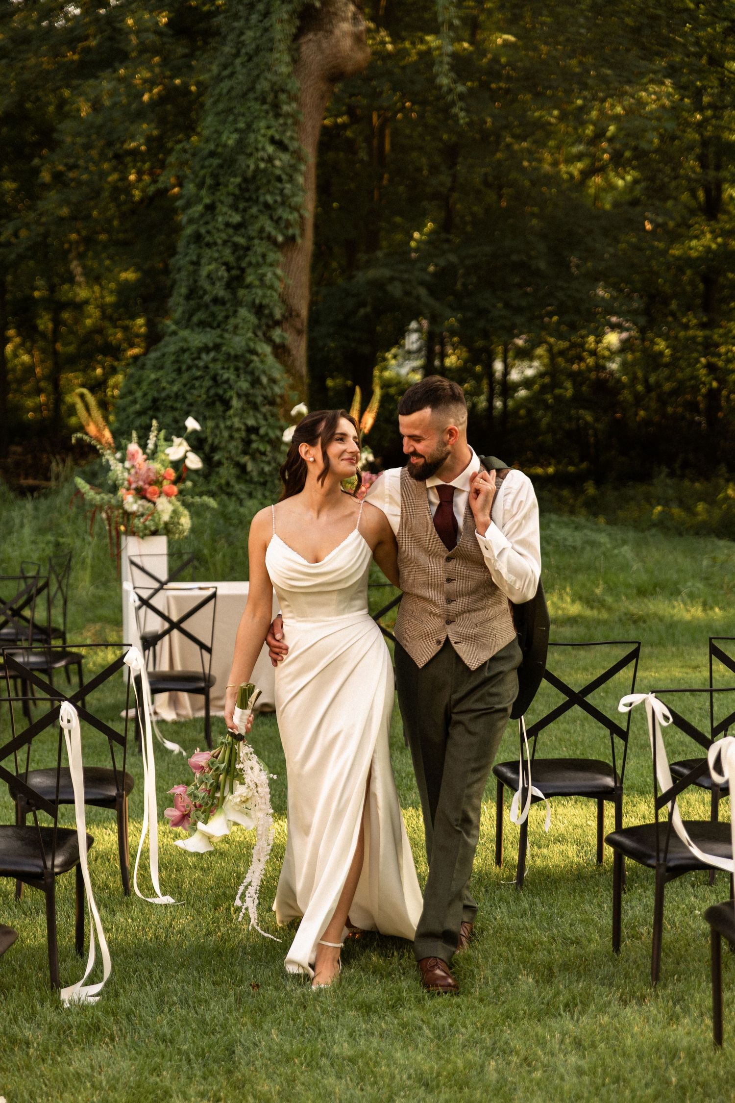 A newlywed couple walks joyfully on grass between guest chairs at an outdoor garden wedding ceremony.