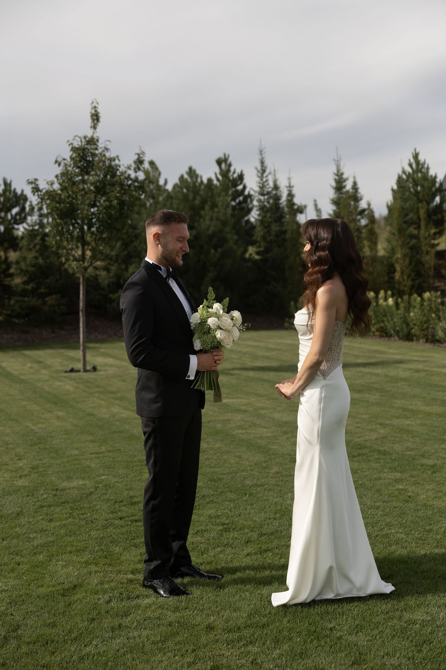 Bride in white gown and groom in black tuxedo share a romantic first look on a lush green lawn.