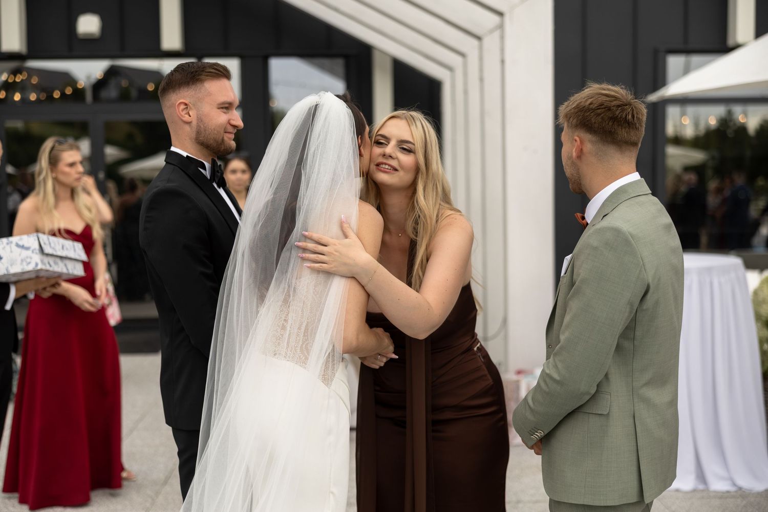 Wedding ceremony moment with bride, groom in tuxedo, bridesmaid in brown dress, and groomsman in sage suit outdoors.