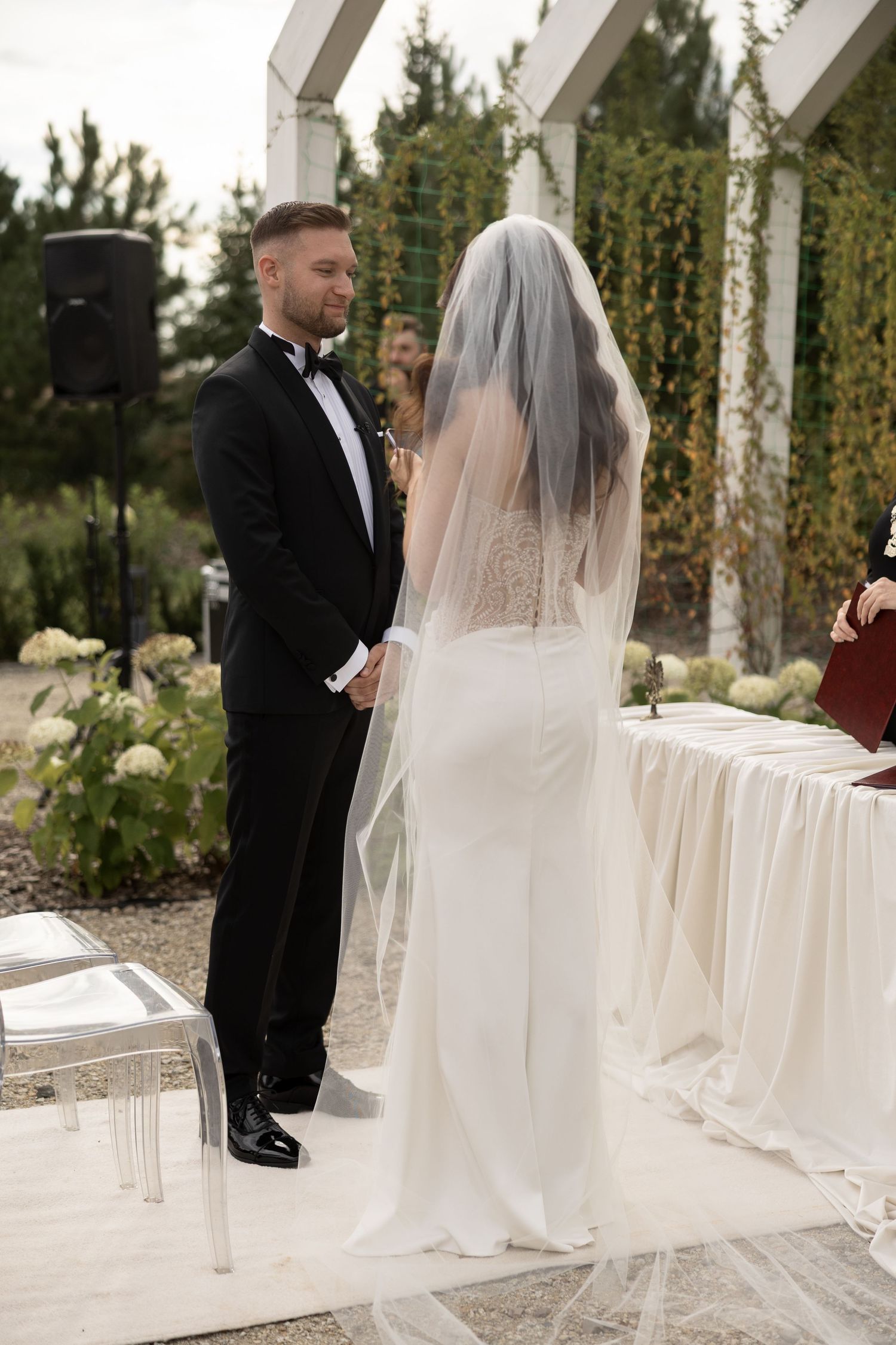 Bride in white gown and veil stands at outdoor wedding altar facing groom in black tuxedo during ceremony.
