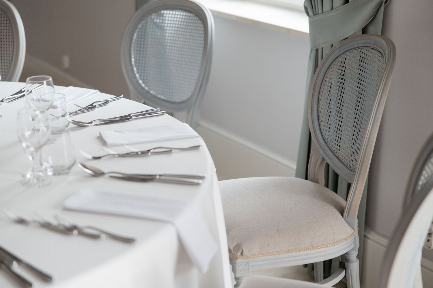 Elegant white dining table set with silverware, glasses, and napkins beside ornate gray French-style chairs in a bright restaurant.