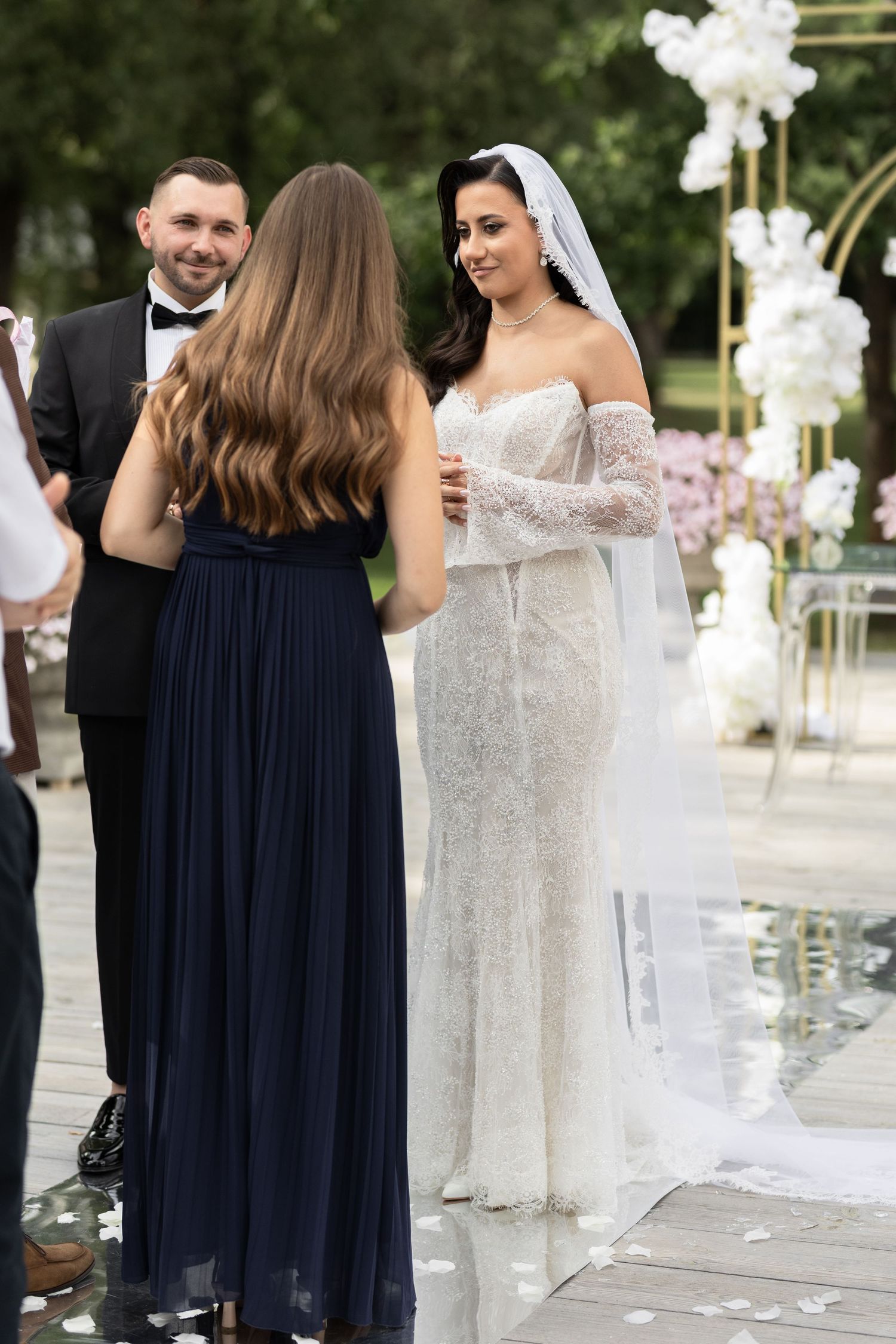 Bride in white lace gown and veil speaks with bridesmaid in navy dress during outdoor wedding ceremony.