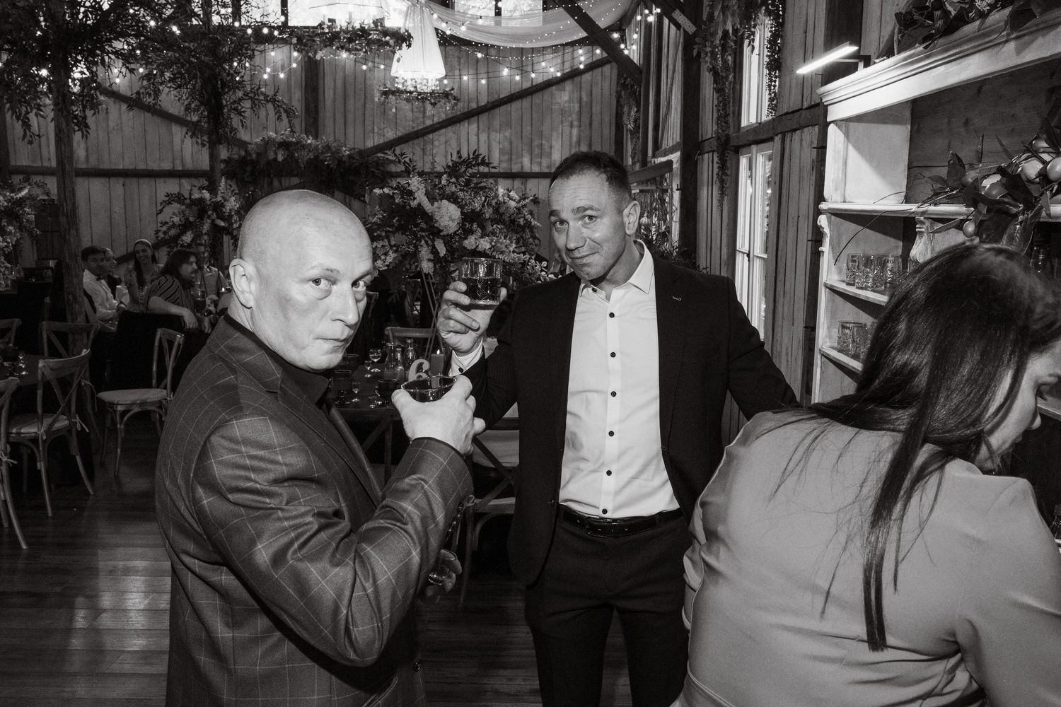 A monochrome photo shows people socializing at an indoor gathering, one person gesturing with a drink in their hand.