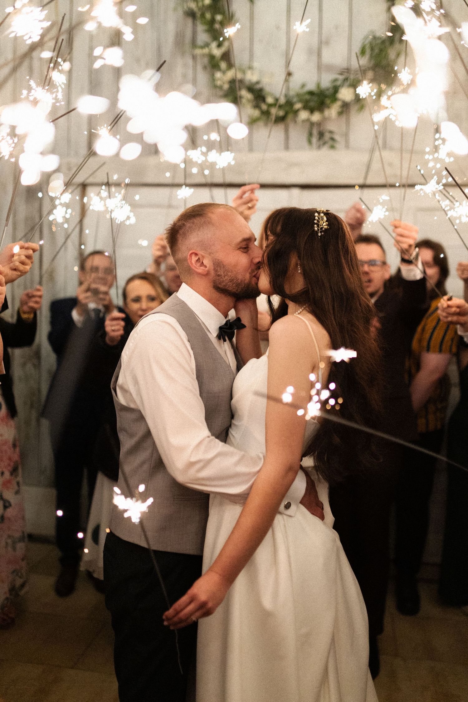 A newly married couple kiss at their indoor reception while guests celebrate with sparklers around them in a romantic scene.