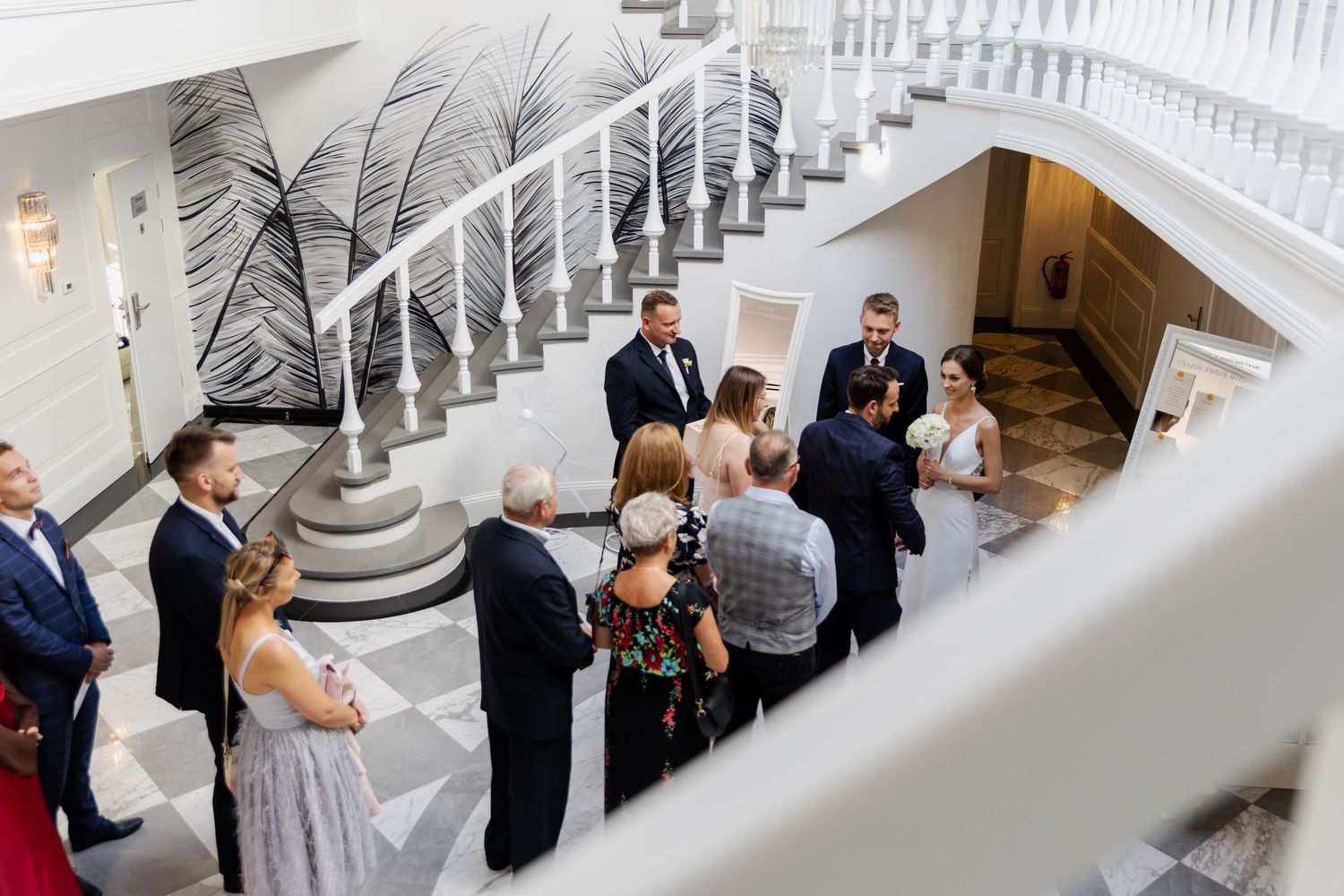 A wedding ceremony gathering viewed from above through white staircase railings in an elegant venue space with palm decor.