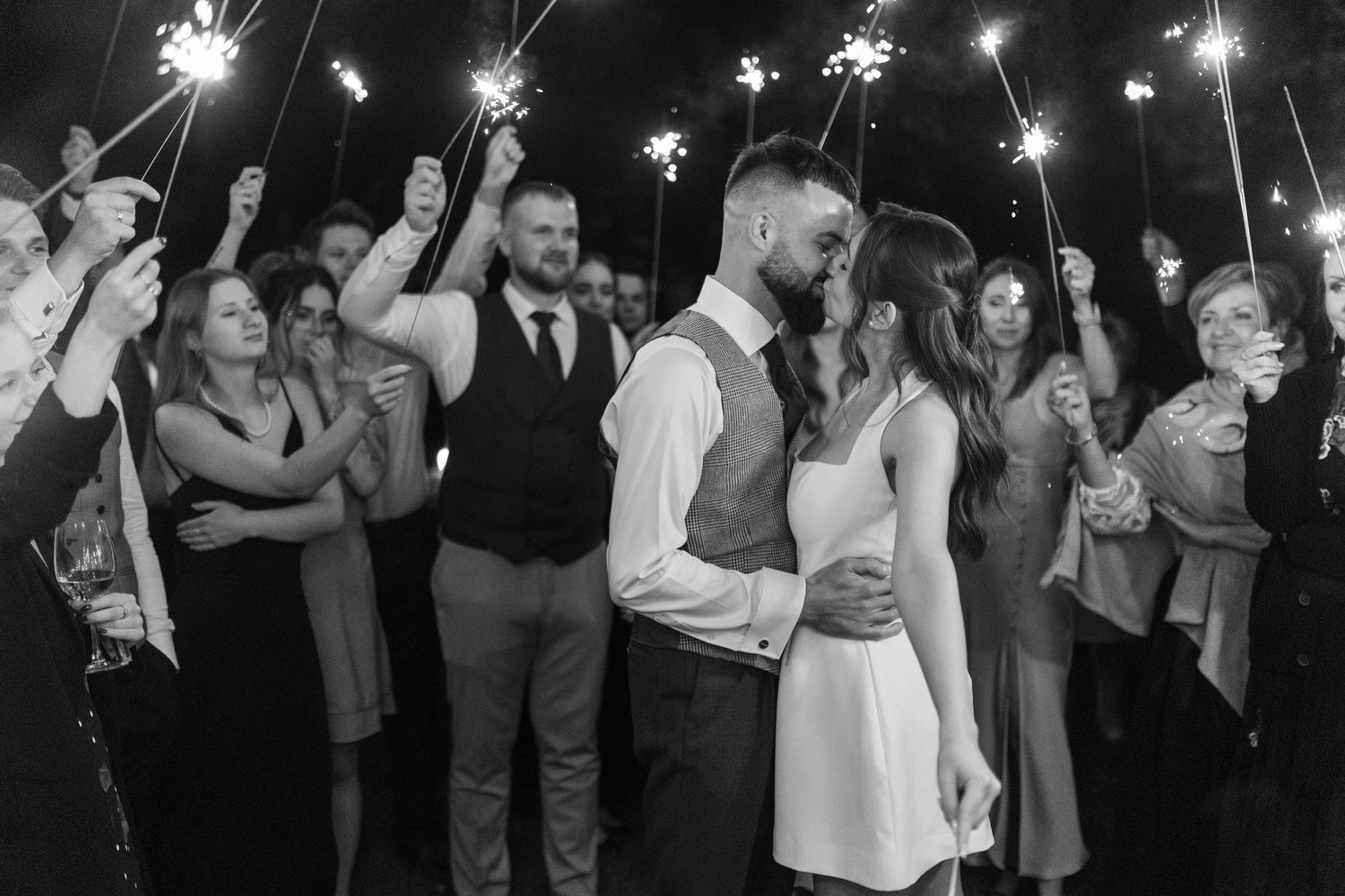 A romantic couple shares a kiss at night surrounded by guests holding glowing sparklers at a wedding celebration.