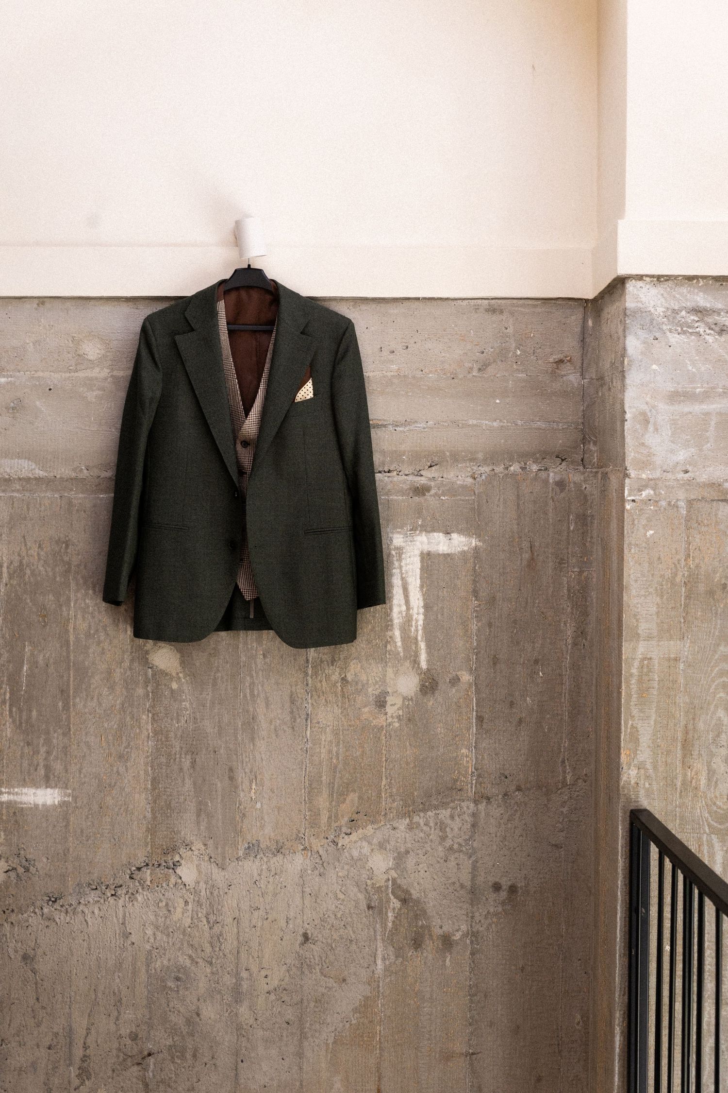 Dark suit jacket hanging alone on concrete wall with visible weathering and metal railing in minimalist artistic setting.