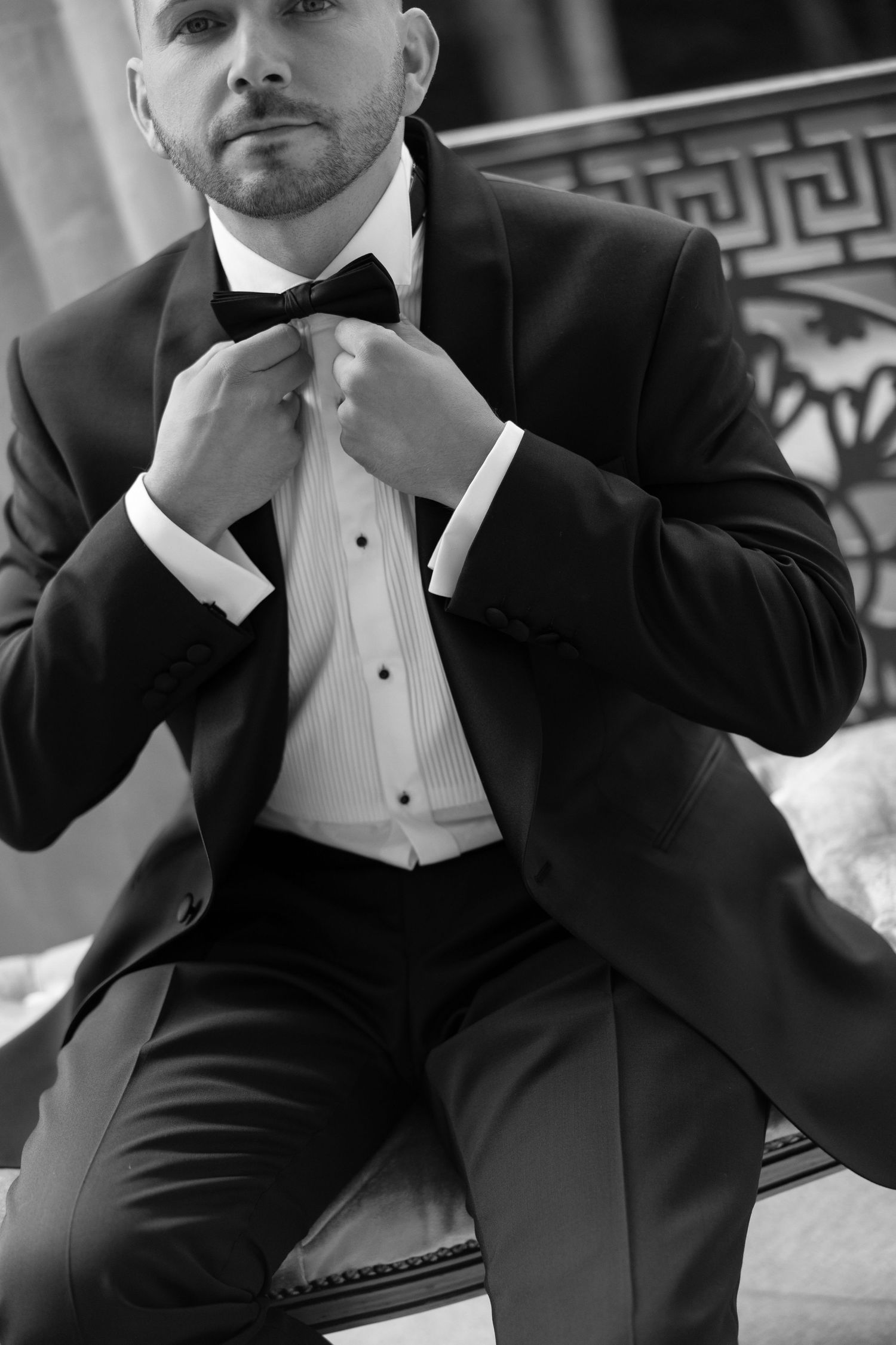 Black and white photo of a well-dressed man in a tuxedo adjusting his bow tie while seated.