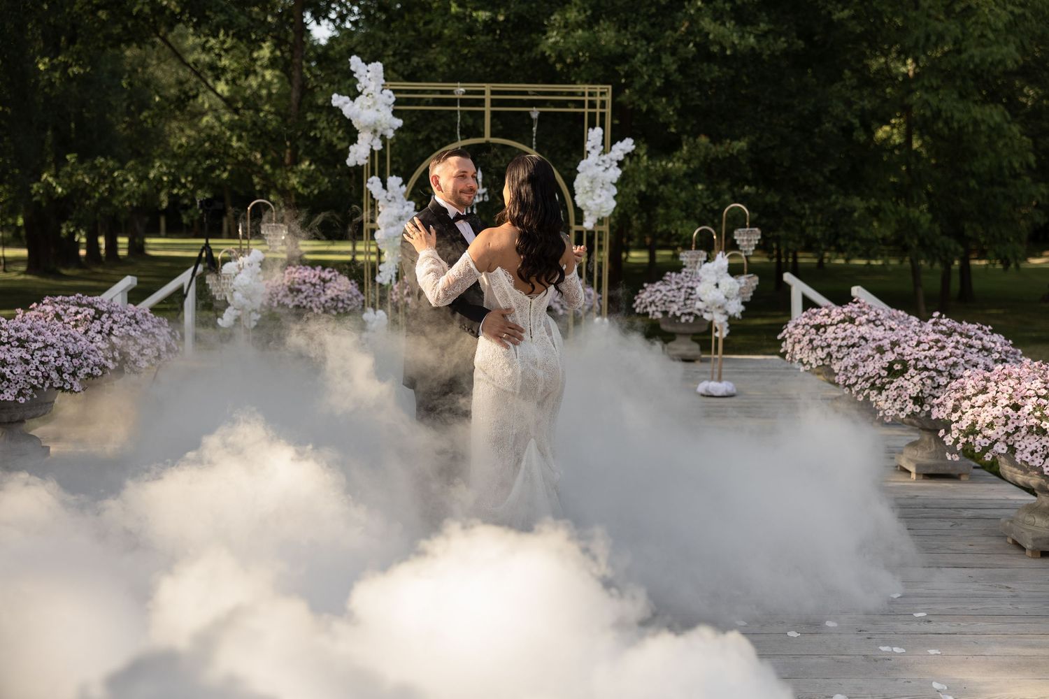 Bride and groom embracing at outdoor wedding ceremony surrounded by dramatic white fog clouds and floral arch decorations.