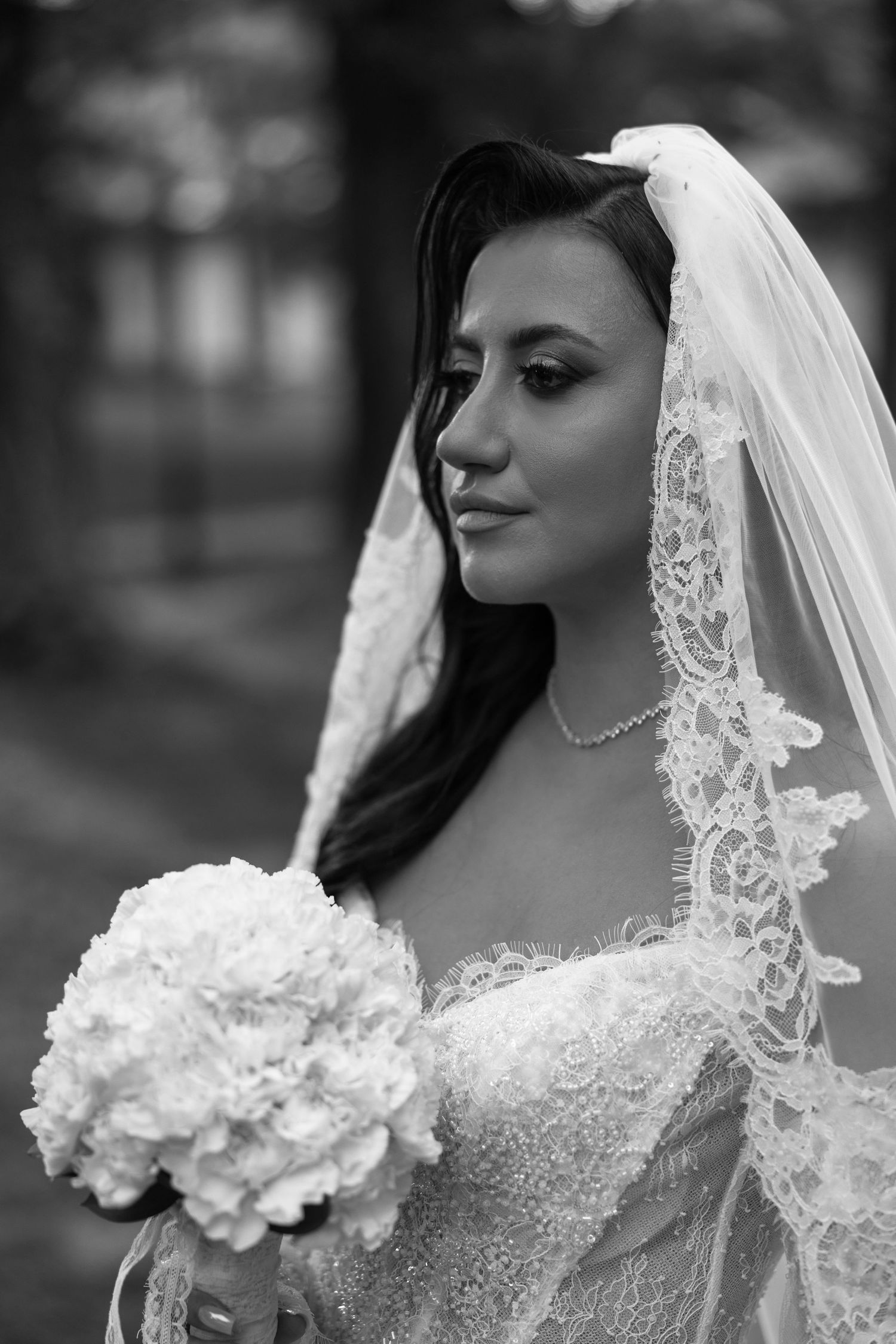 Black and white portrait of a bride in lace wedding dress and veil holding a white floral bouquet outdoors.