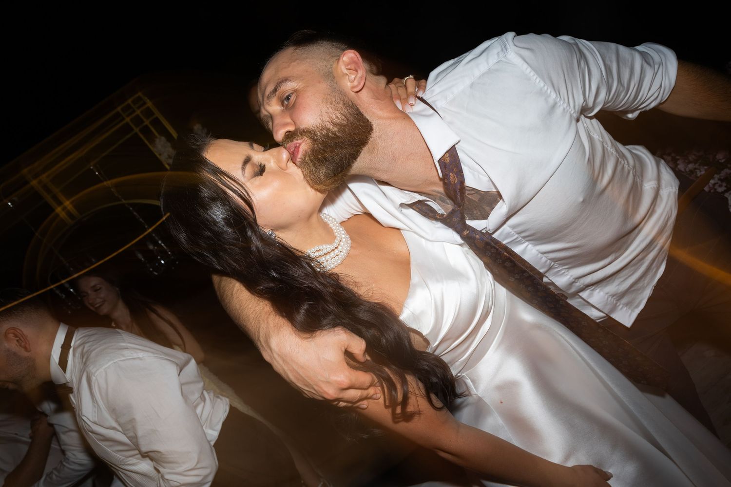 Couple sharing a romantic kiss during their wedding reception, groom in white shirt, bride in satin gown.