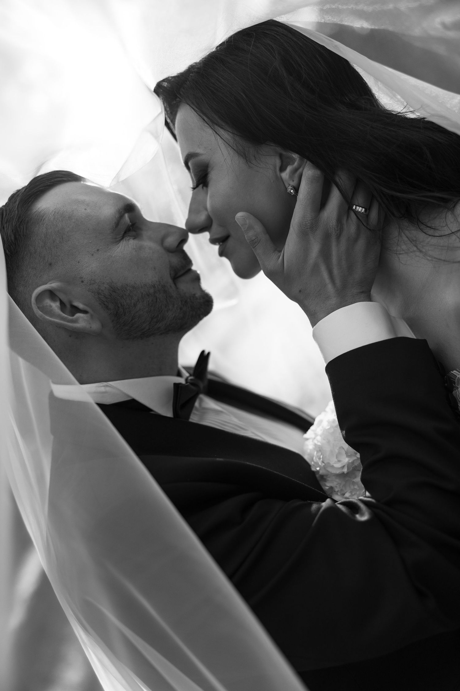 Black and white photo of a bride and groom sharing a romantic moment, nearly kissing under a flowing veil.