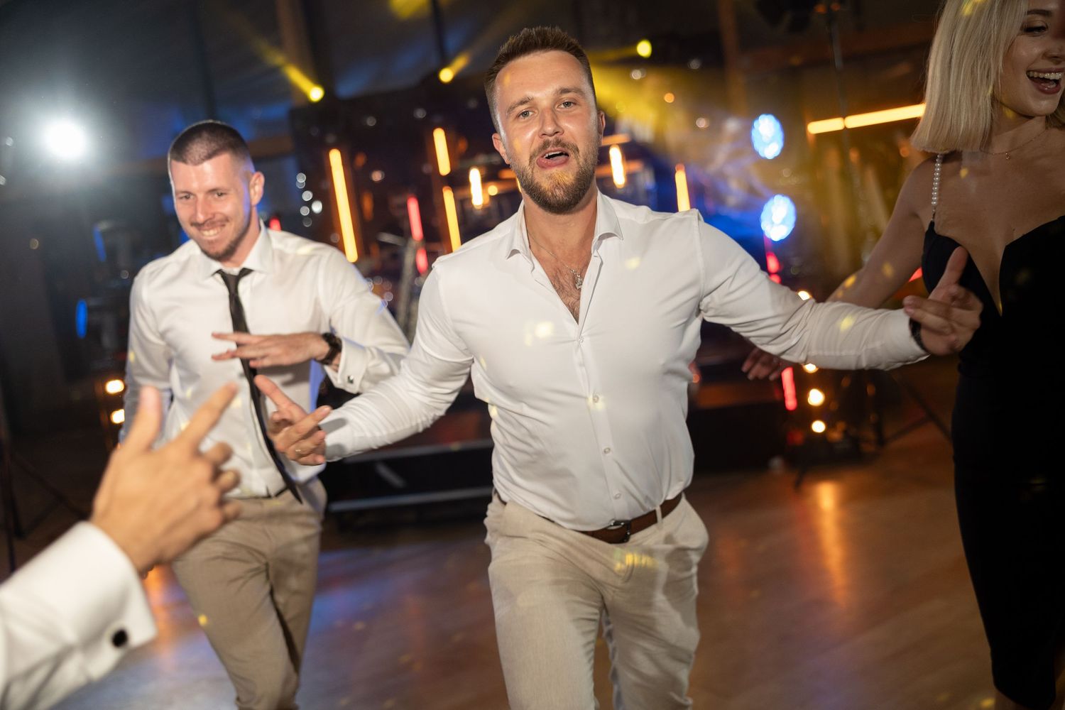Two men dancing energetically at a party or wedding reception on a dance floor with festive lighting.