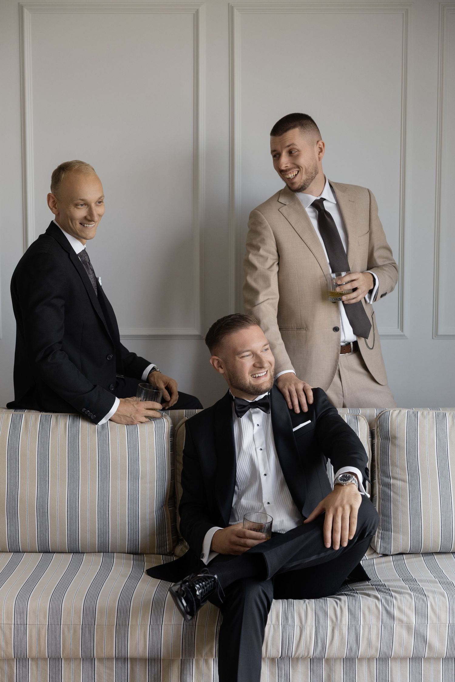 Three well-dressed men in formal suits socializing on a striped sofa in an elegant room.