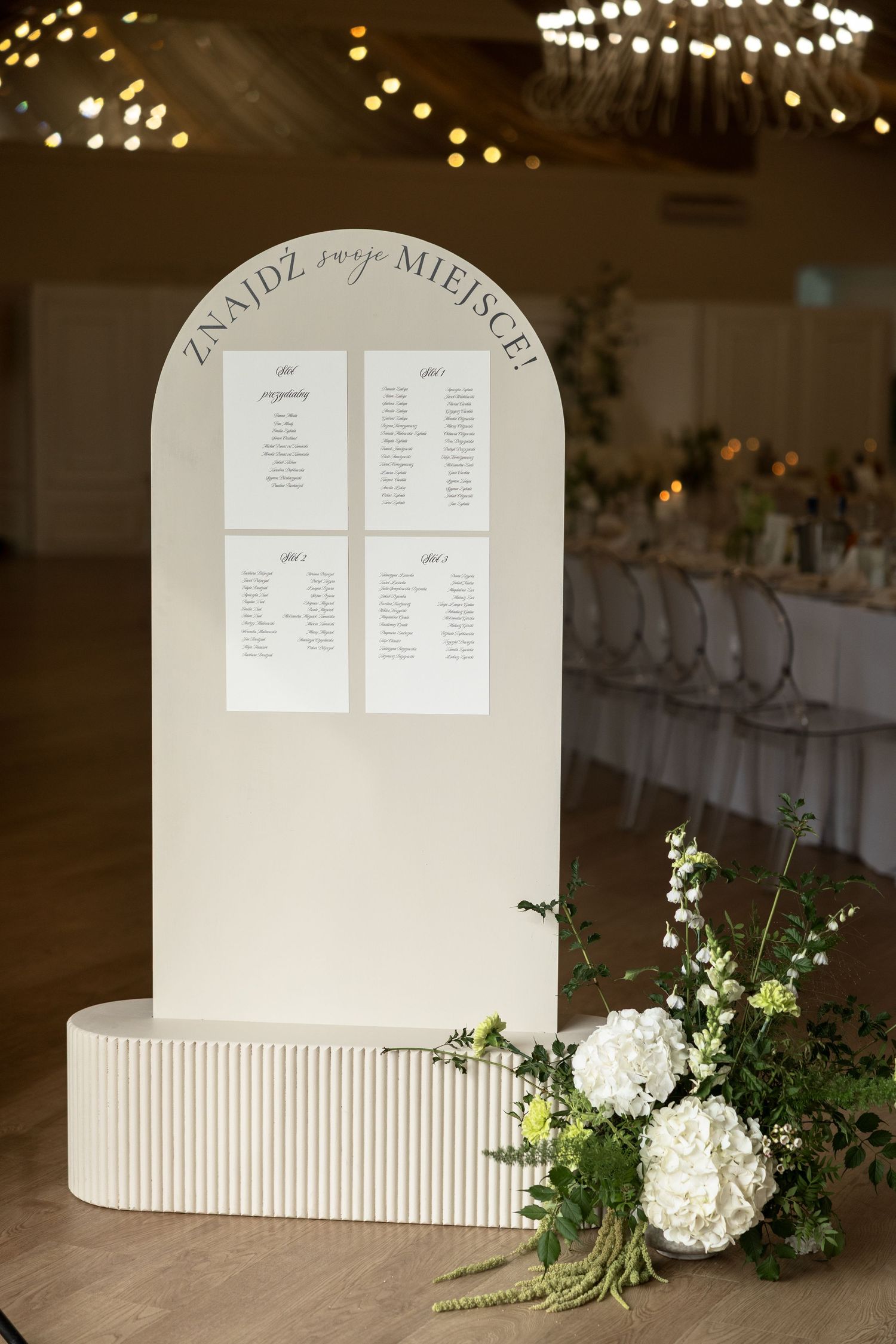 Elegant wedding seating chart display on arch stand with white floral arrangement and table assignments in a reception hall.