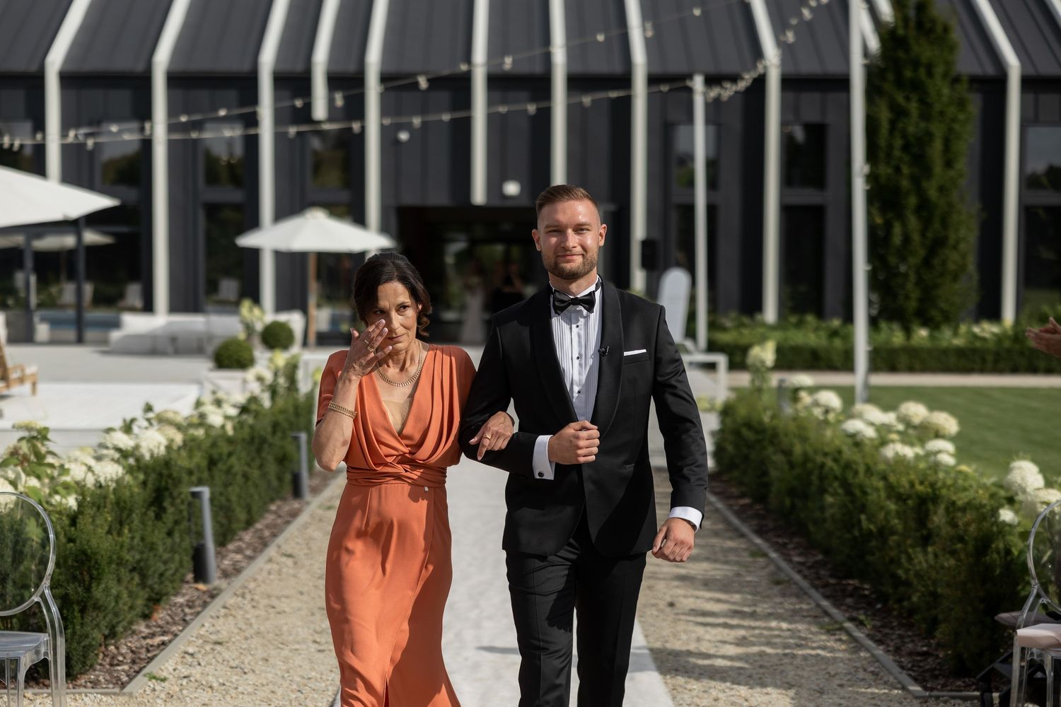 Elegant couple walking outdoors at a venue, woman in orange gown and man in black tuxedo with bow tie.