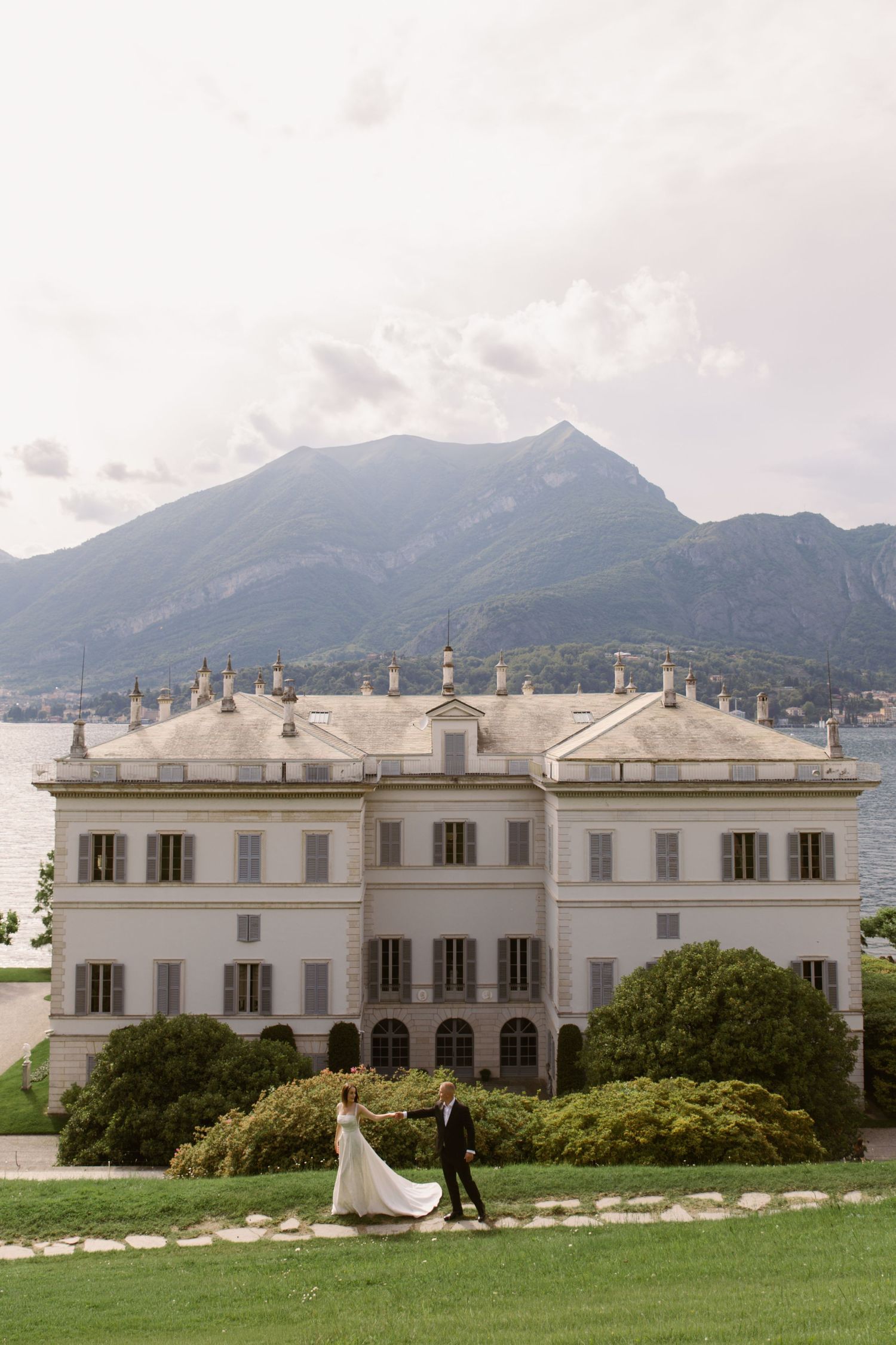 Bride and groom walking in front of elegant Italian villa on Lake Como with mountain backdrop.
