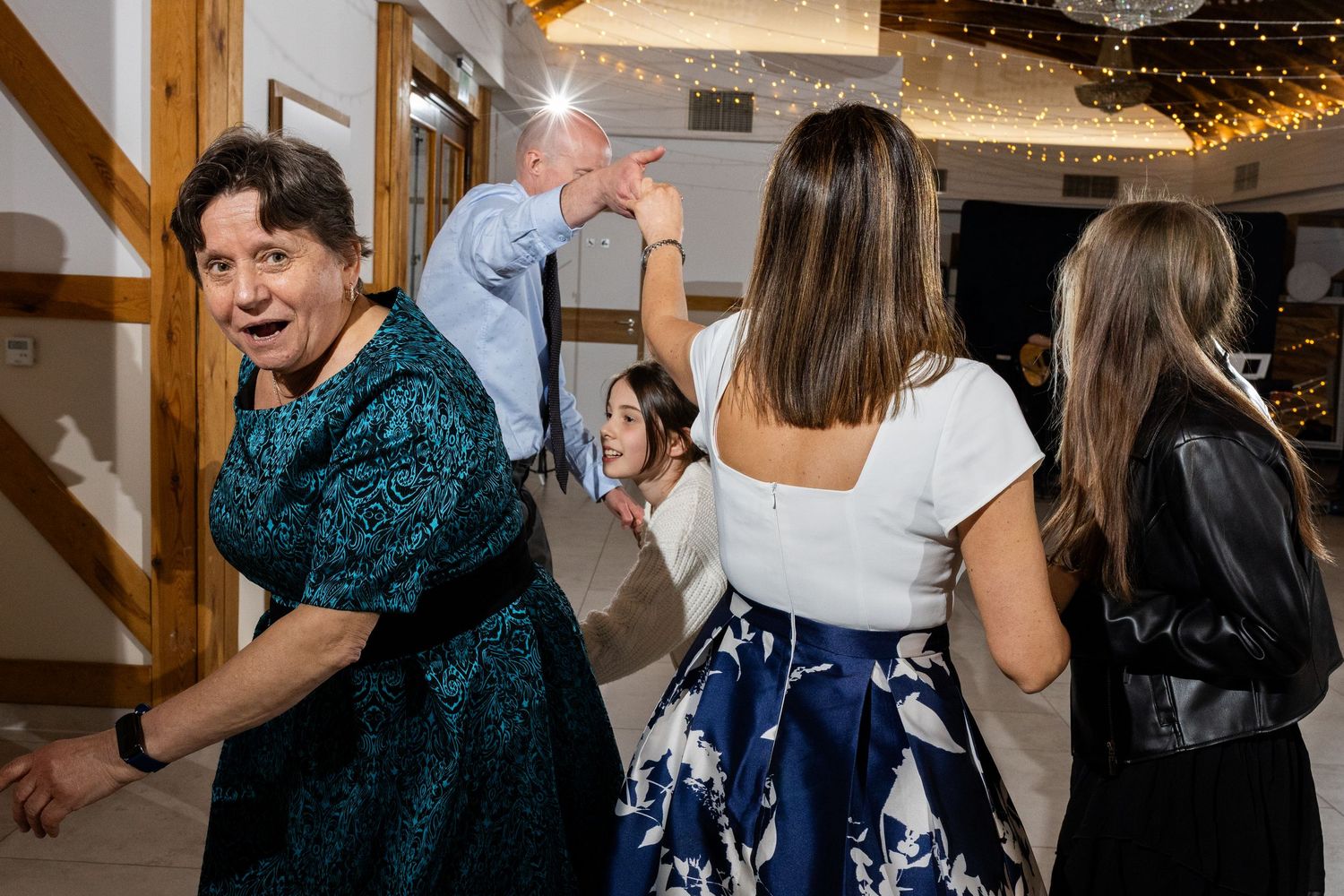 Guests dancing energetically at a wedding reception under twinkling lights in a rustic venue.