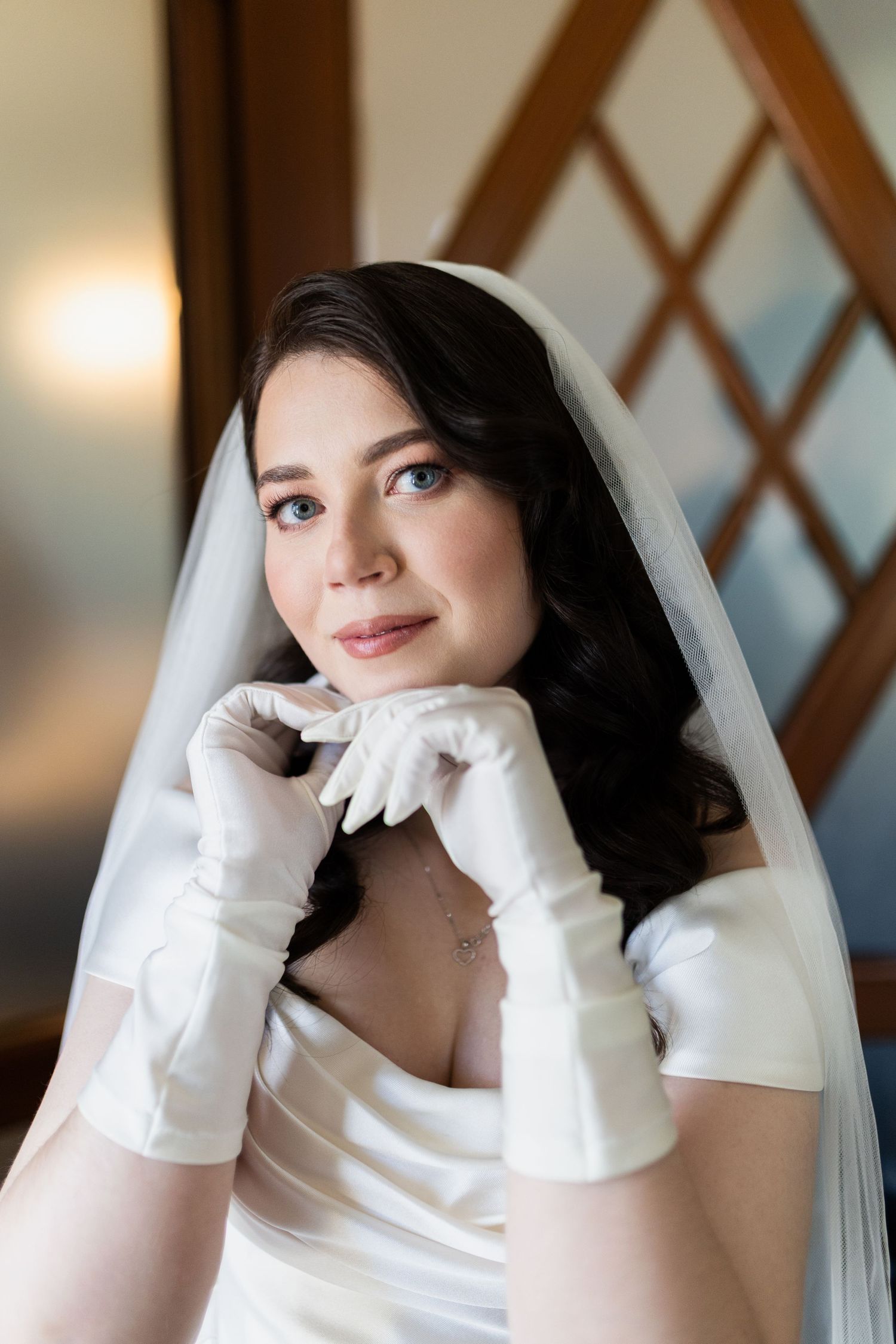 A bride wearing a classic white wedding gown with long satin gloves poses elegantly against a latticed background.