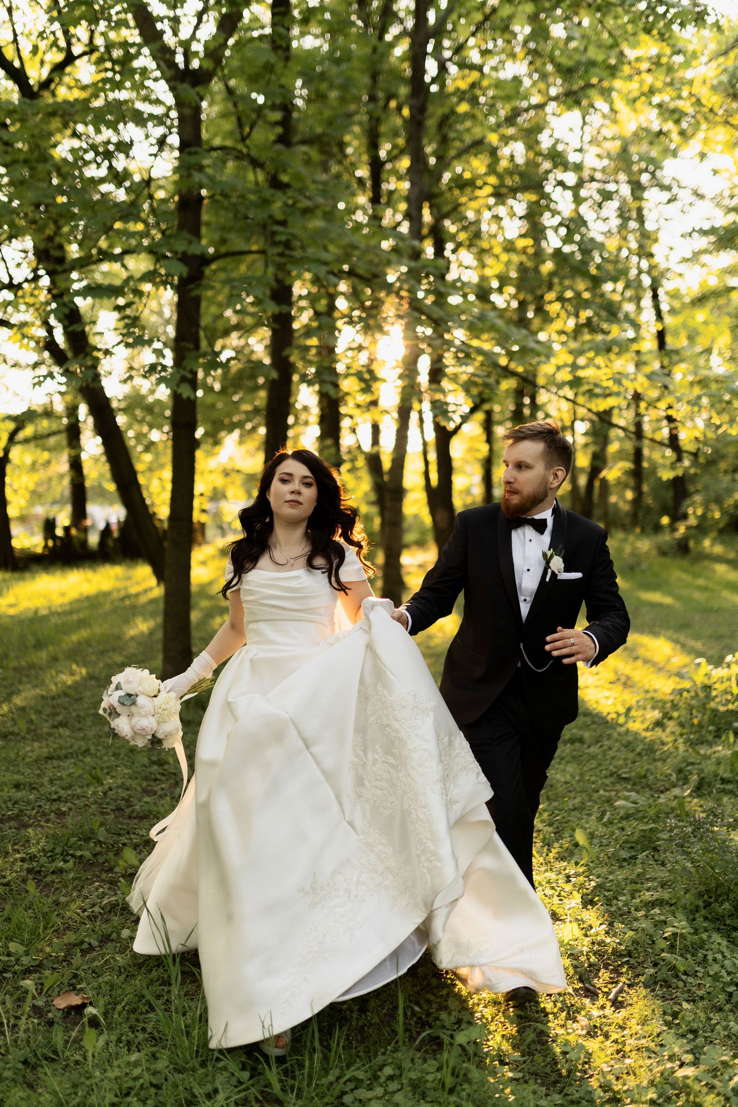 A couple in wedding attire walks through a sunlit forest with golden afternoon light filtering through the trees.