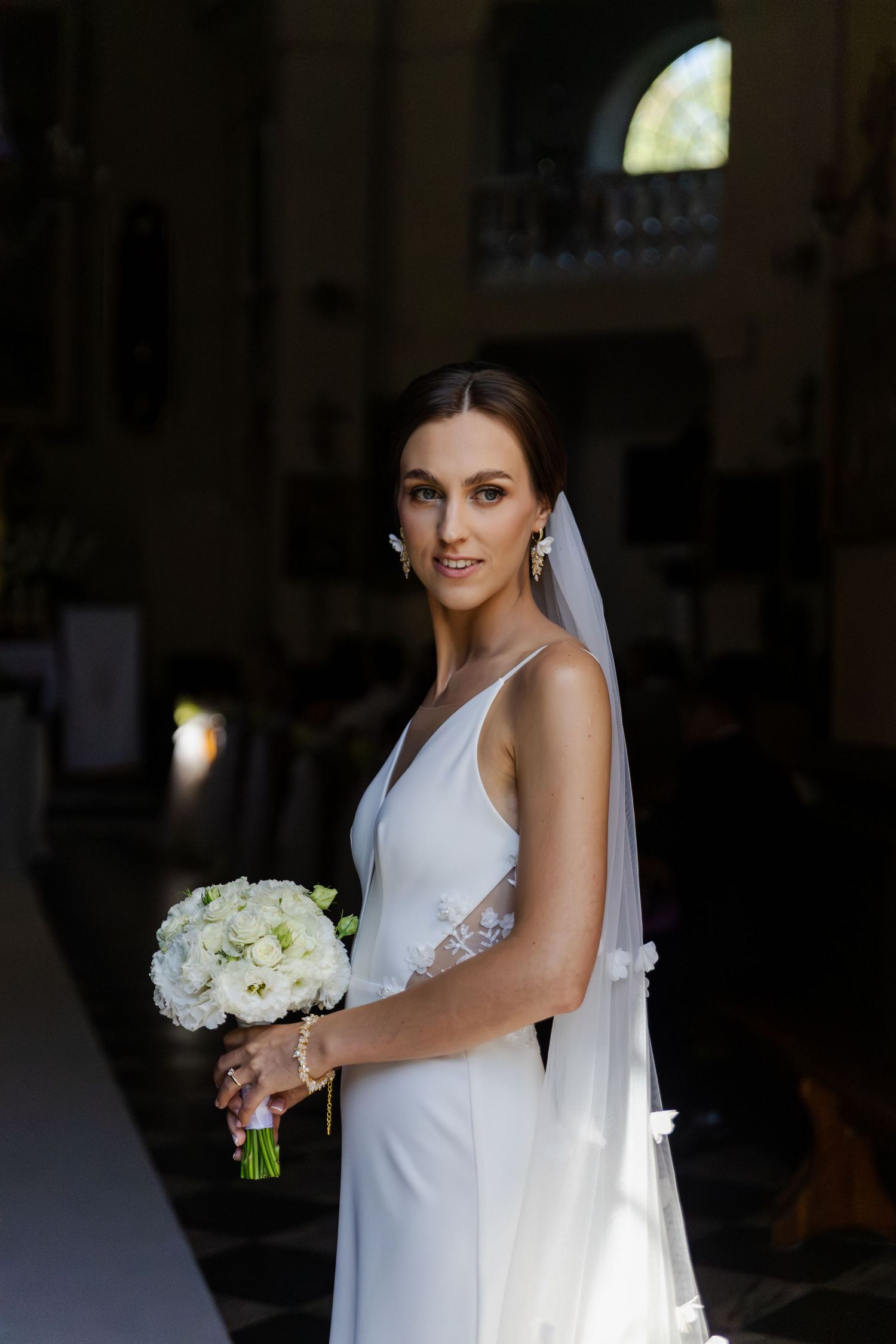 Beautiful bride in sleek white wedding gown holds classic white bouquet in church with natural light streaming through window.